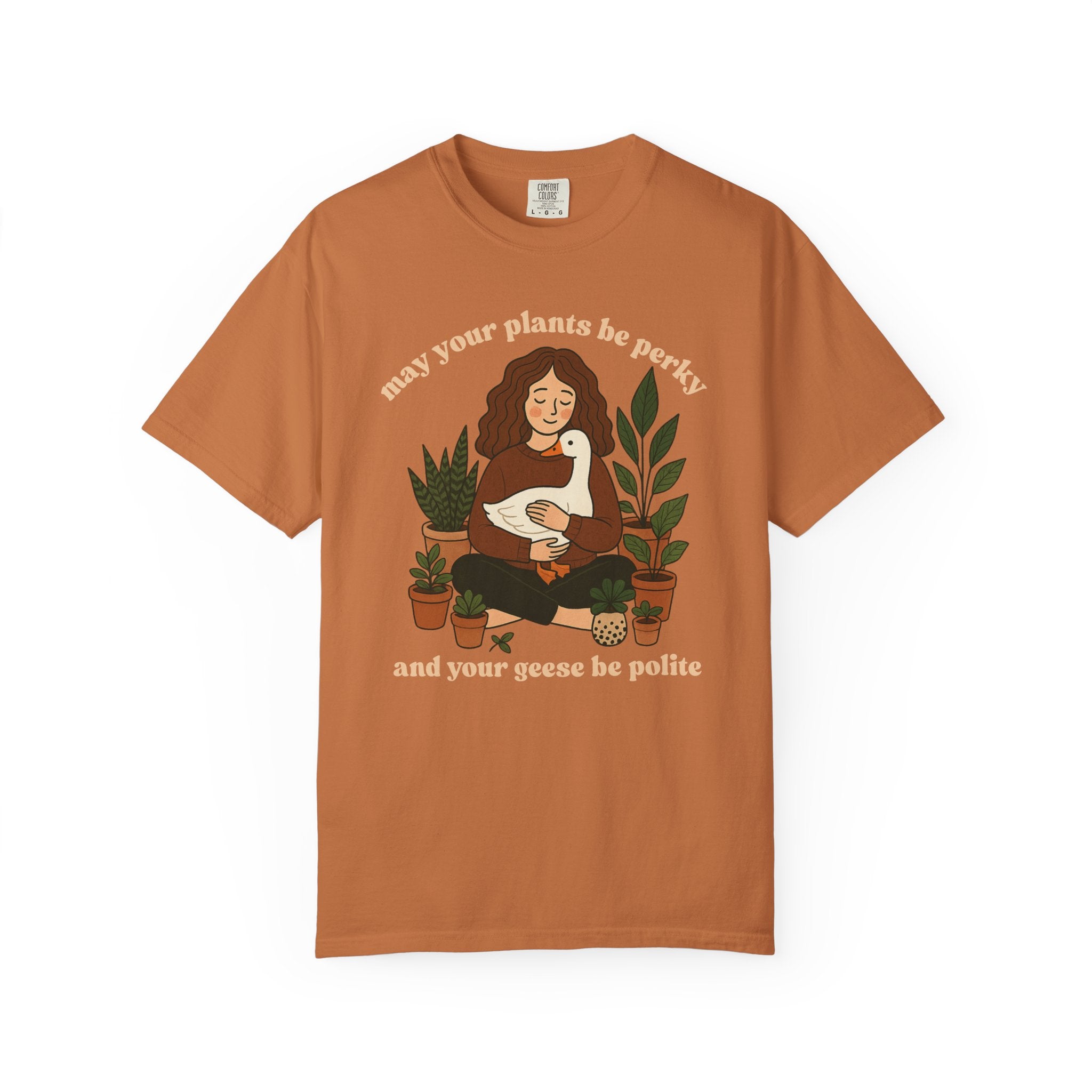 A cozy woman holding a goose surrounded by potted plants with the text “May your plants be perky and your geese be polite” printed on a Comfort Colors cottagecore shirt.