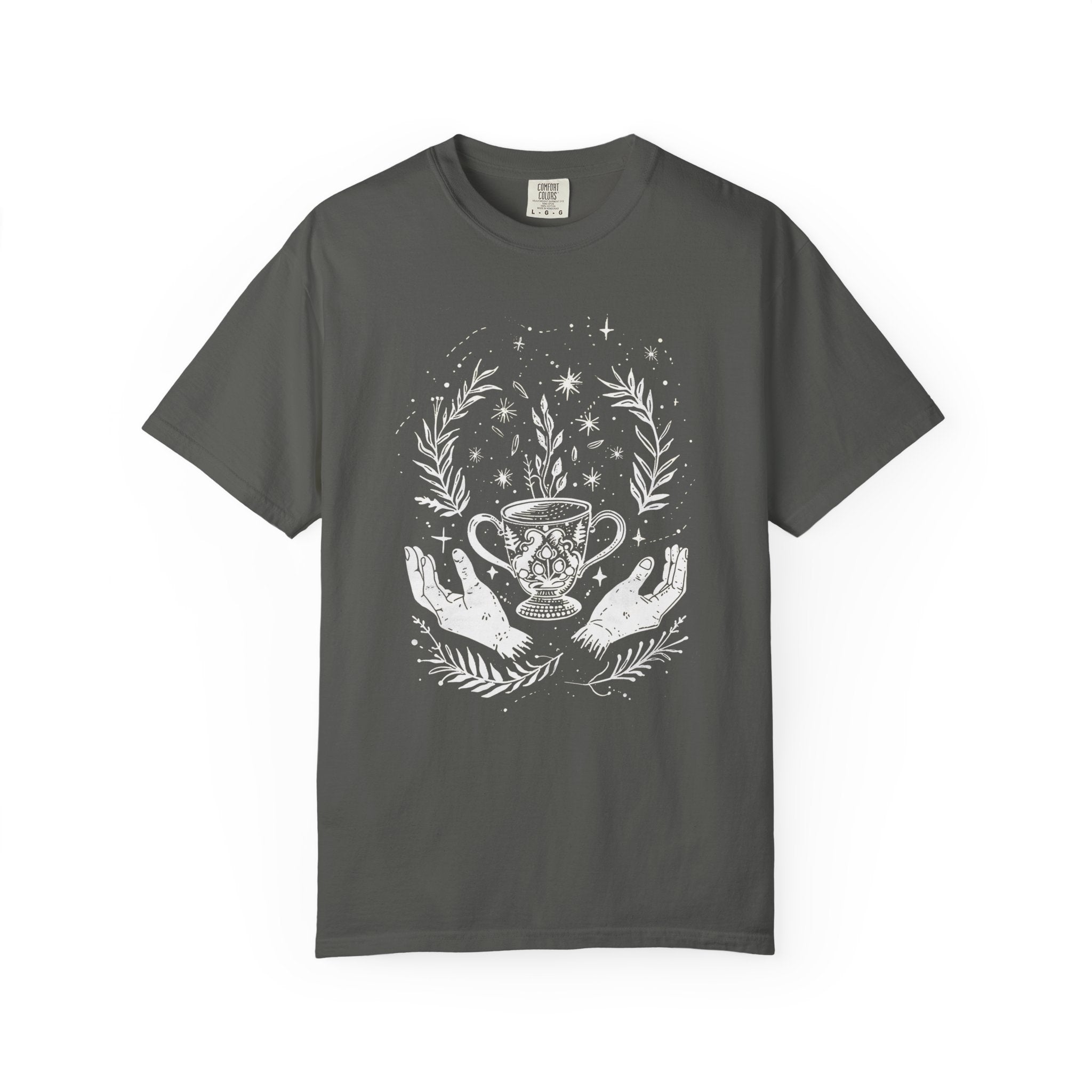 Black Comfort Colors style t-shirt featuring a mystical tea chalice held between open hands, surrounded by stars, botanical leaves, and celestial witchy elements in a cottagecore tea witch aesthetic.