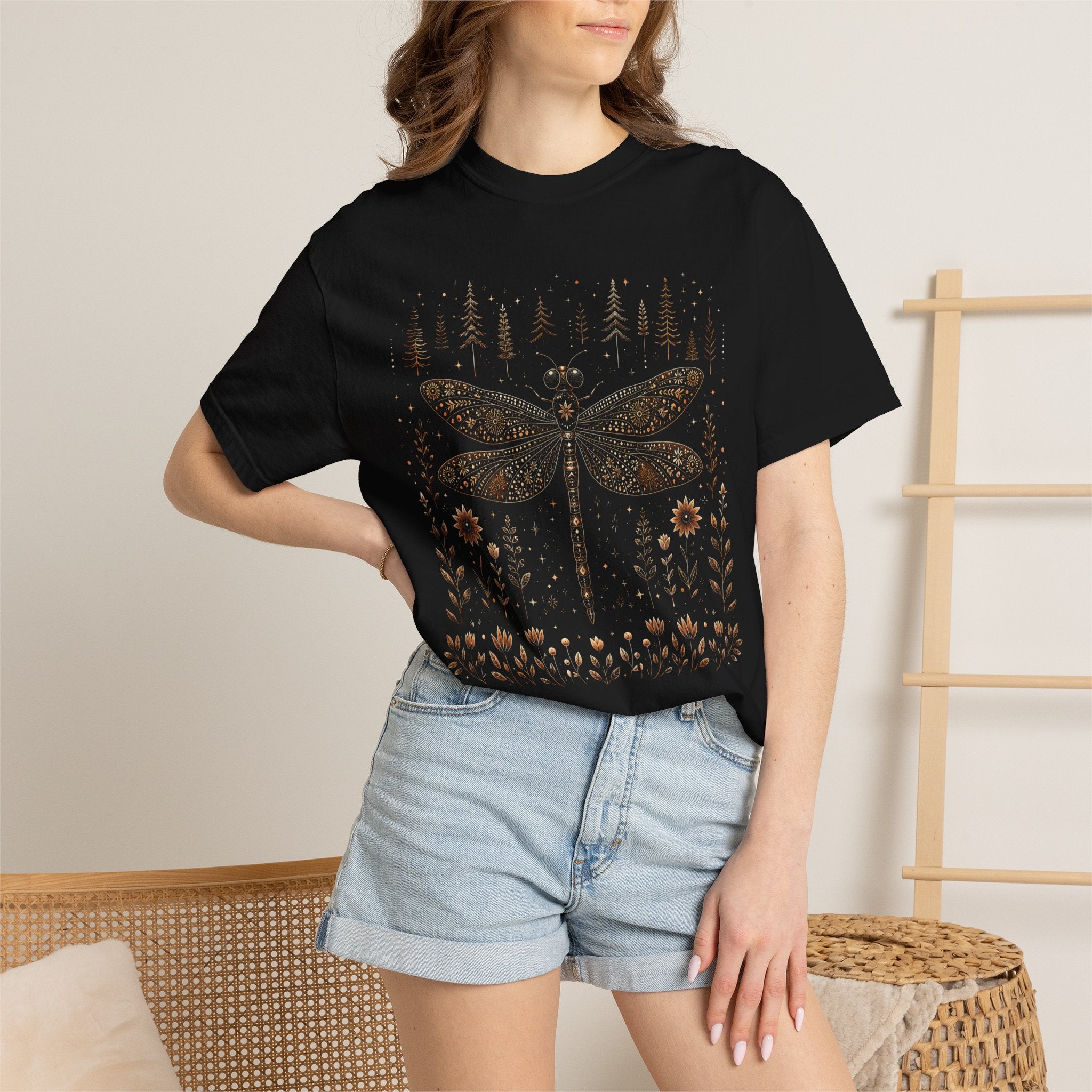 Whimsical vintage-style dragonfly illustration with intricate floral patterns on its wings, surrounded by wildflowers, pine trees, stars, and celestial details in warm bronze and cream tones against a black background.