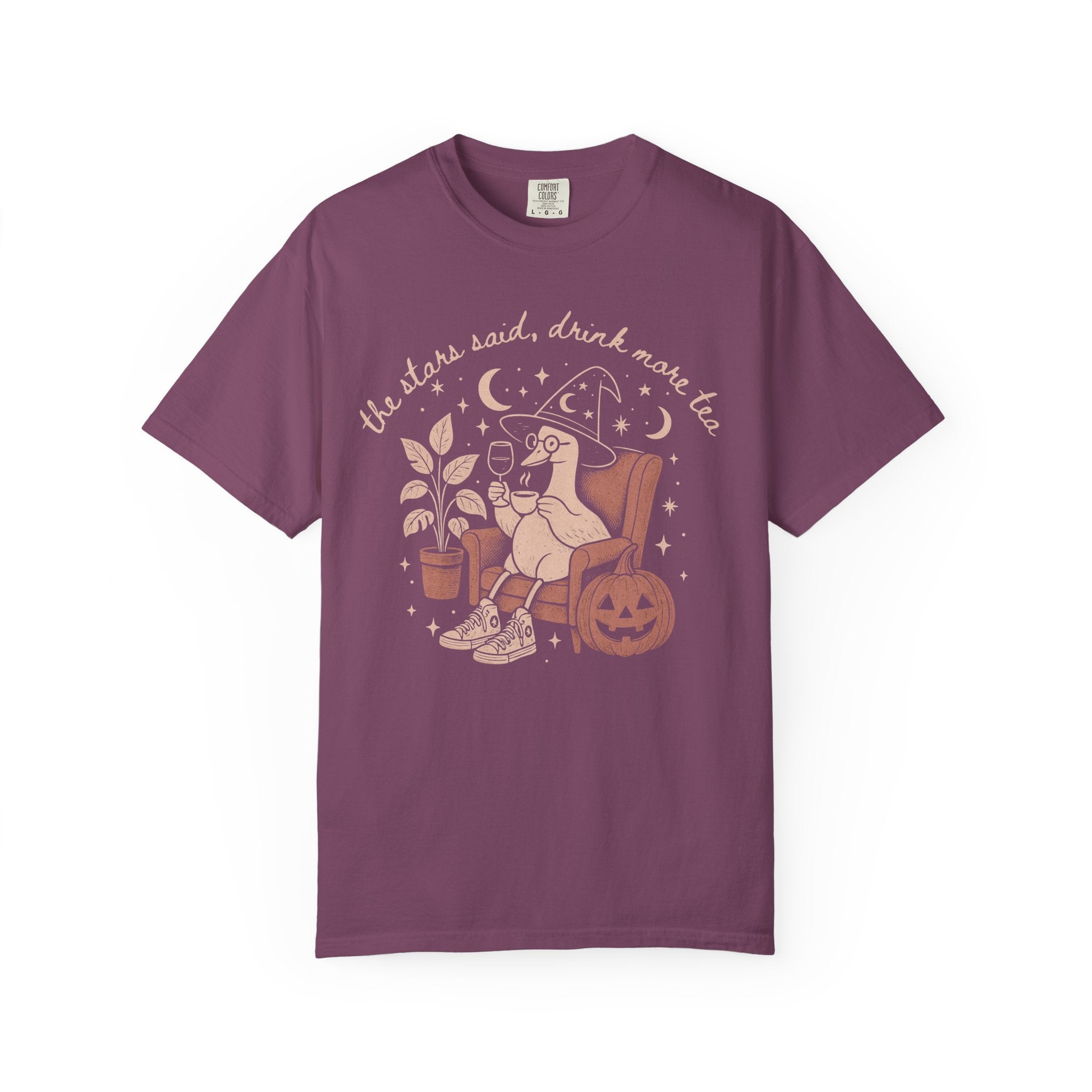 Comfort Colors graphic t-shirt featuring a cozy witch goose sitting in an armchair holding tea and wine beside a pumpkin with stars and moons and the quote "The Stars Said, Drink More Tea."