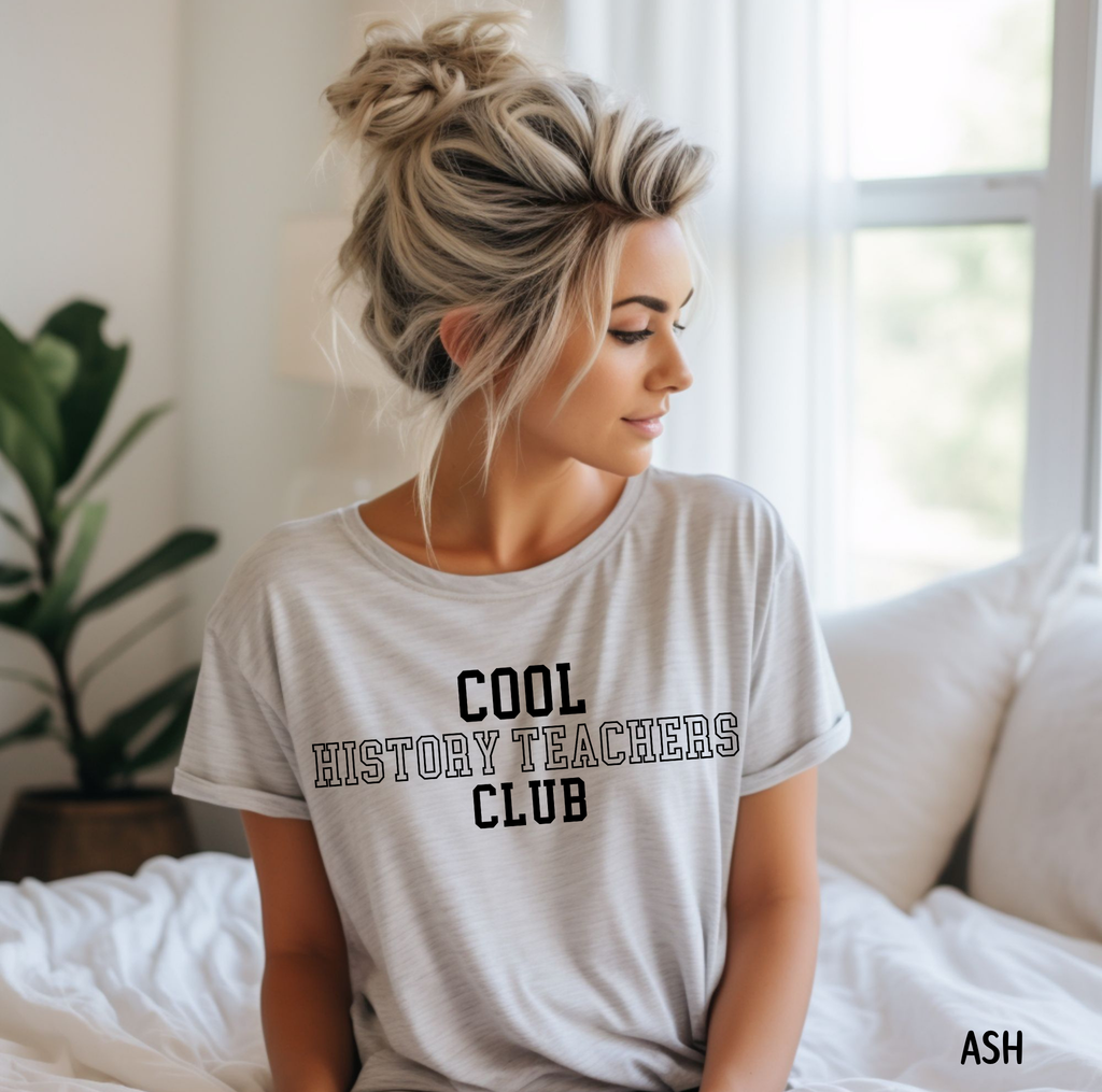 A woman with blonde hair in a messy bun sits on a bed wearing a light gray t-shirt with the text 'COOL HISTORY TEACHERS CLUB' printed in black. The background shows a bright bedroom with white bedding, a window, and a green plant.