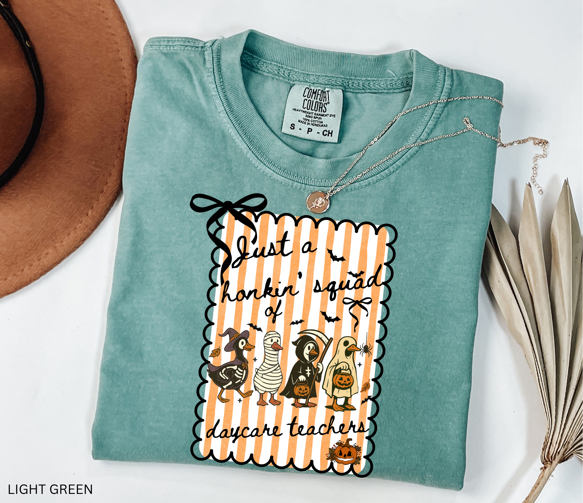 Halloween daycare teacher shirt featuring four geese dressed in spooky costumes (witch, mummy, reaper, ghost) with pumpkins, bats, and candy on orange striped background. Perfect group Halloween tee for daycare teachers.