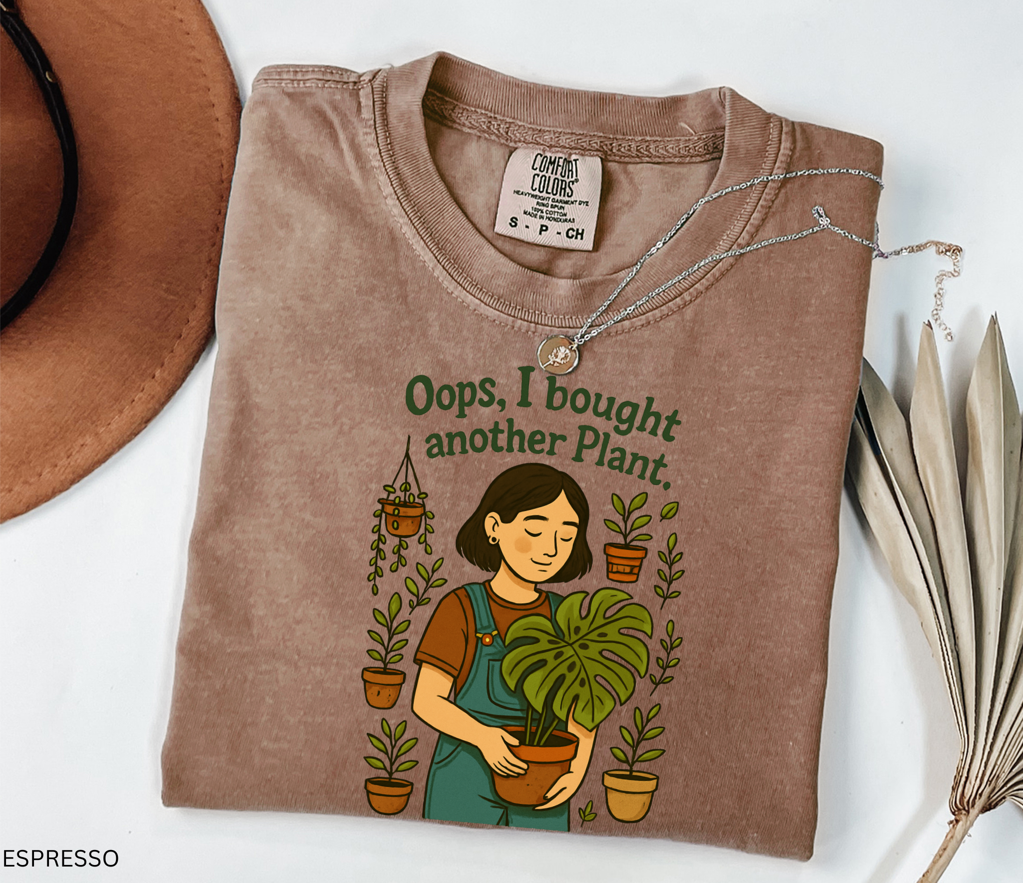 Gardening shirt with a woman in overalls holding a monstera plant, surrounded by potted plants, with the phrase “Oops, I Bought Another Plant.”