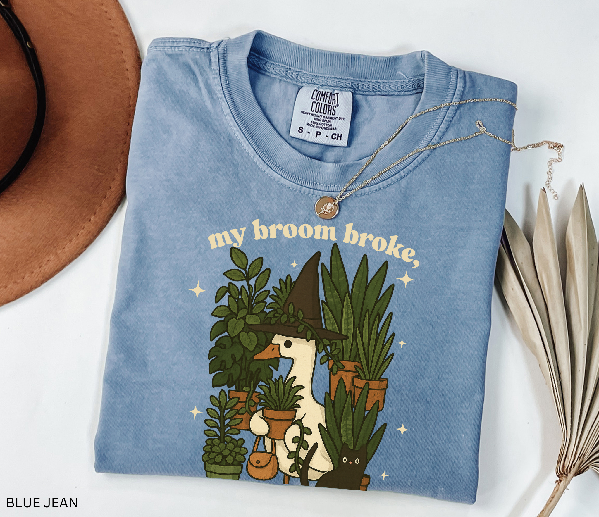 Witchy goose holding plants with a black cat surrounded by greenery — “My broom broke, now I tend to plants” text on cozy Comfort Colors tee.