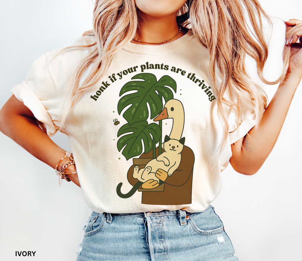 Goose holding a cat beside a monstera plant with the text “Honk If Your Plants Are Thriving” on a Comfort Colors shirt.