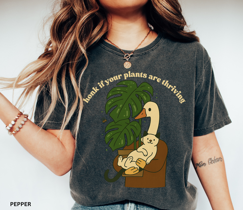 Goose holding a cat beside a monstera plant with the text “Honk If Your Plants Are Thriving” on a Comfort Colors shirt.