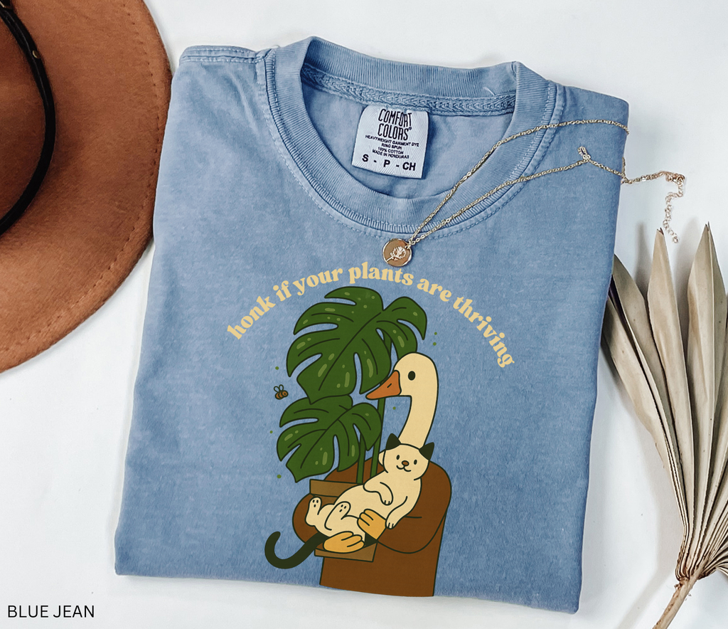 Goose holding a cat beside a monstera plant with the text “Honk If Your Plants Are Thriving” on a Comfort Colors shirt.