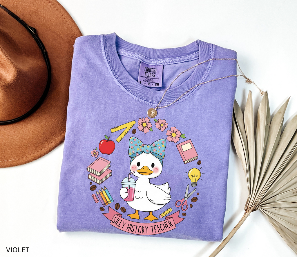 Funny history teacher shirt with a goose wearing a bow, surrounded by school doodles like books, pencils, apple, and scissors. Reads ‘Silly History Teacher.