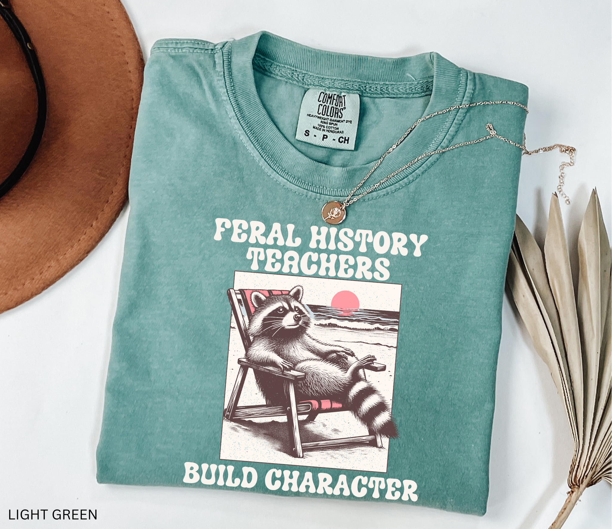 Funny history teacher shirt with a raccoon lounging in a beach chair and the phrase ‘Feral History – Teachers Build Character.’ Humorous social studies teacher tee
