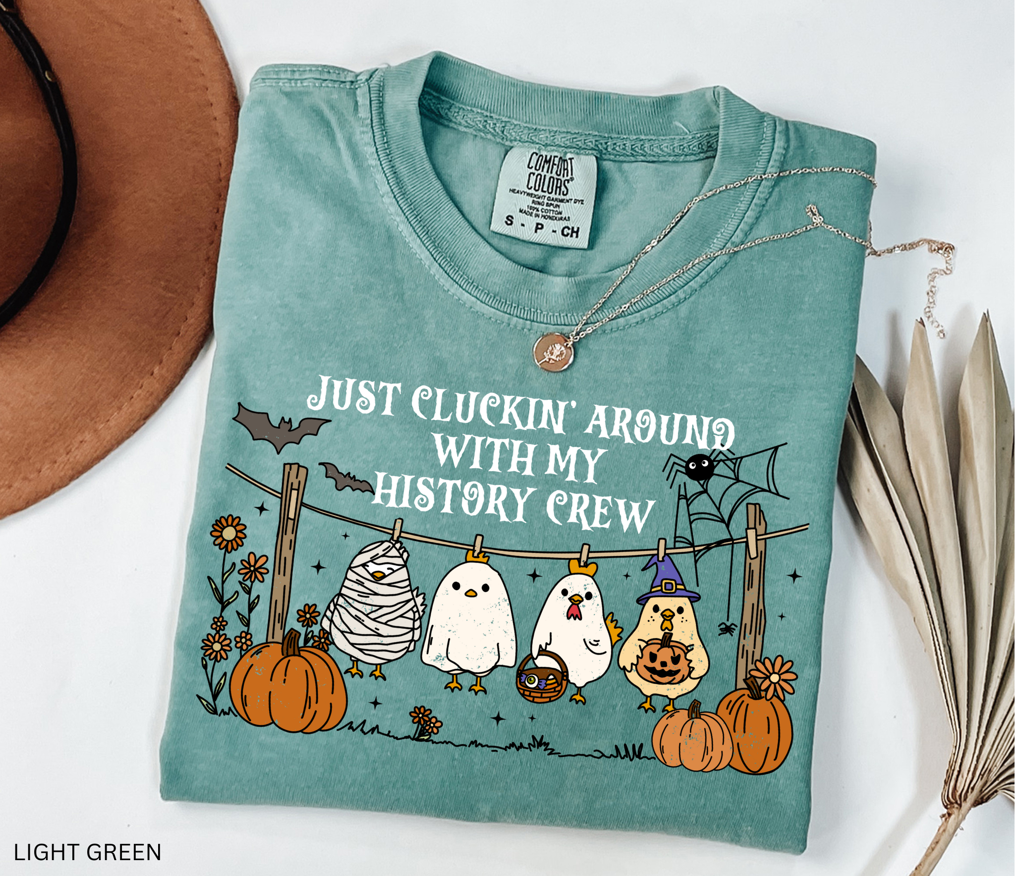 Halloween history teacher shirt with chickens in costumes on a clothesline, pumpkins, bats, and the phrase ‘Just cluckin’ around with my history crew.’ Funny group teacher shirt.