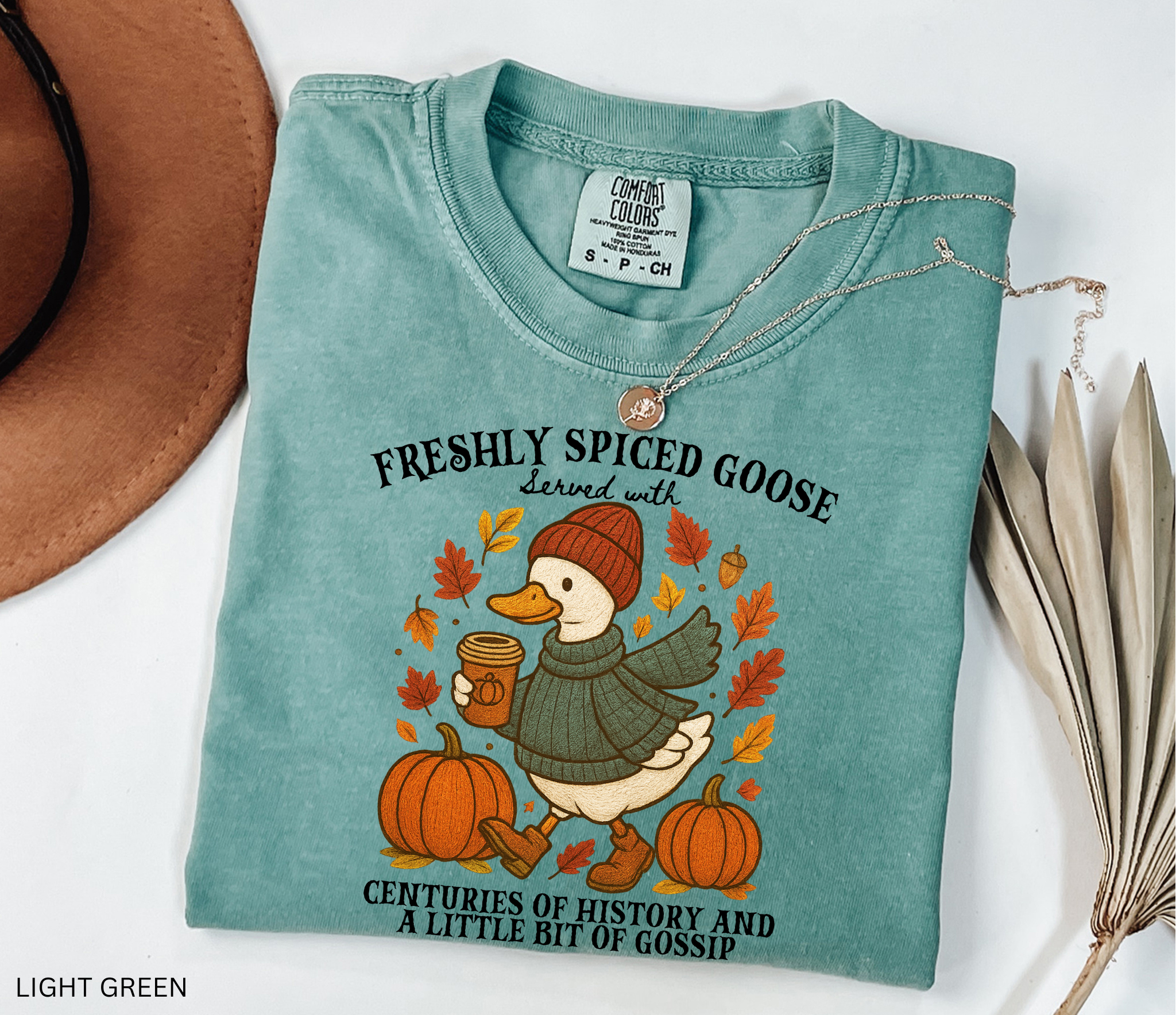 Cozy fall shirt design featuring a goose in a sweater and beanie holding pumpkin spice coffee, surrounded by pumpkins and autumn leaves – made for history teachers and professors.