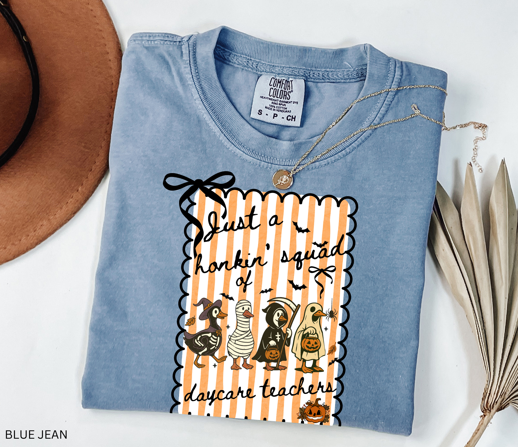 Halloween daycare teacher shirt featuring four geese dressed in spooky costumes (witch, mummy, reaper, ghost) with pumpkins, bats, and candy on orange striped background. Perfect group Halloween tee for daycare teachers.