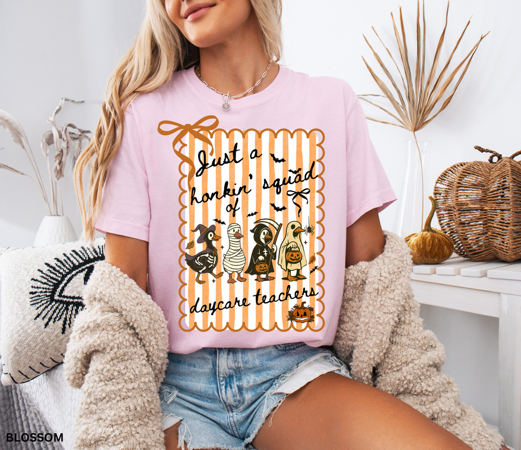Halloween daycare teacher shirt featuring four geese dressed in spooky costumes (witch, mummy, reaper, ghost) with pumpkins, bats, and candy on orange striped background. Perfect group Halloween tee for daycare teachers.