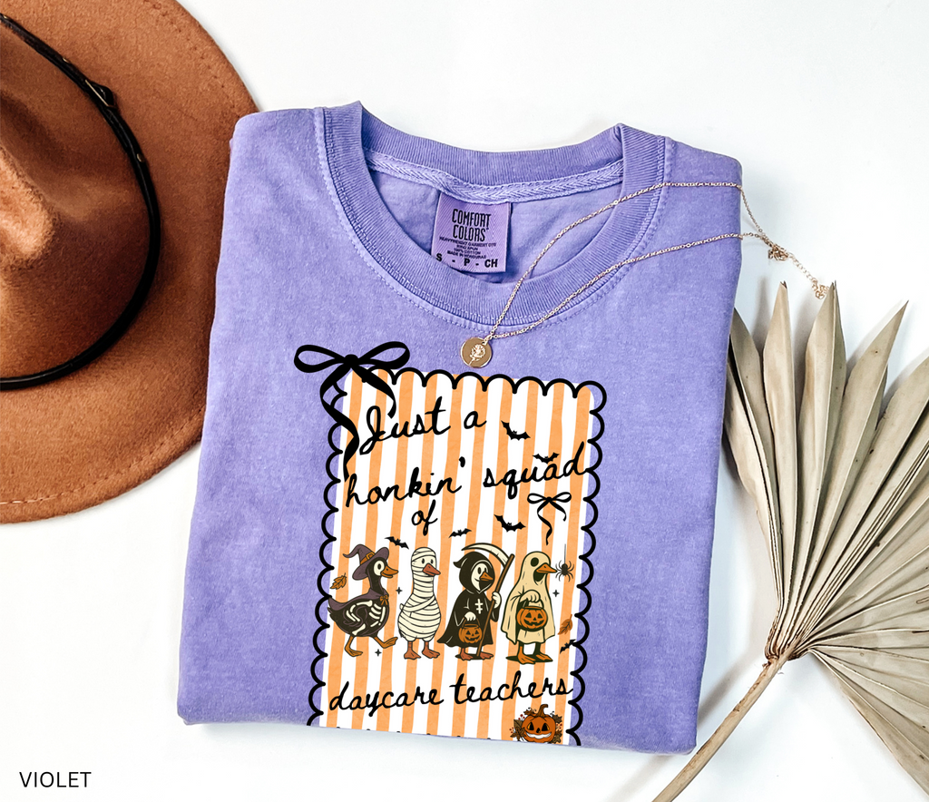 Halloween daycare teacher shirt featuring four geese dressed in spooky costumes (witch, mummy, reaper, ghost) with pumpkins, bats, and candy on orange striped background. Perfect group Halloween tee for daycare teachers.
