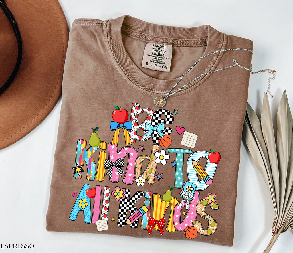 Colorful “Be Kind to All Kinds” teacher shirt with playful letters, apples, pears, bows, flowers, and school-themed accents.