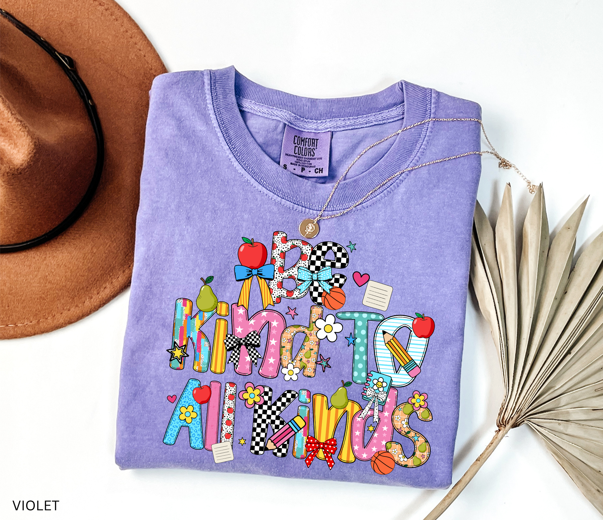 Colorful “Be Kind to All Kinds” teacher shirt with playful letters, apples, pears, bows, flowers, and school-themed accents.