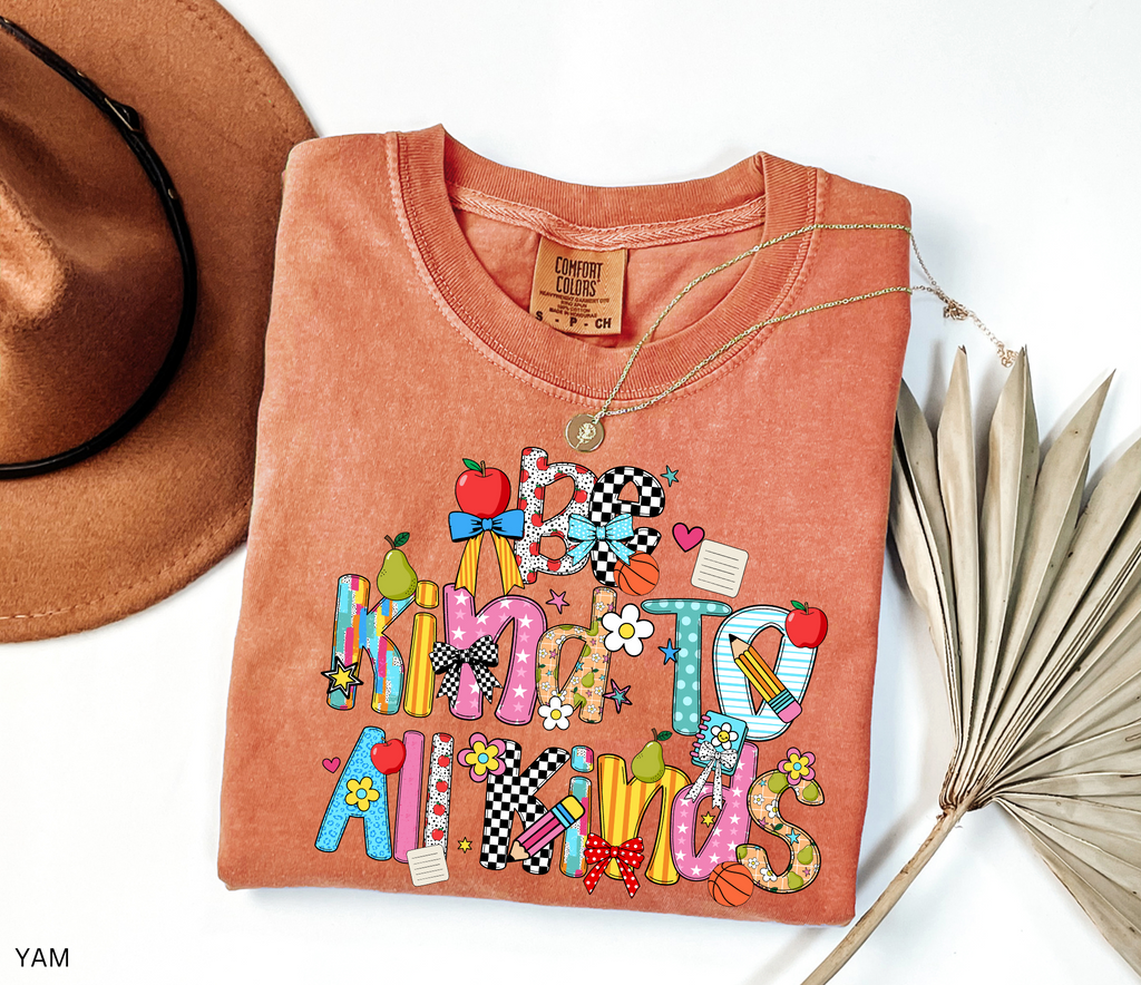 Colorful “Be Kind to All Kinds” teacher shirt with playful letters, apples, pears, bows, flowers, and school-themed accents.
