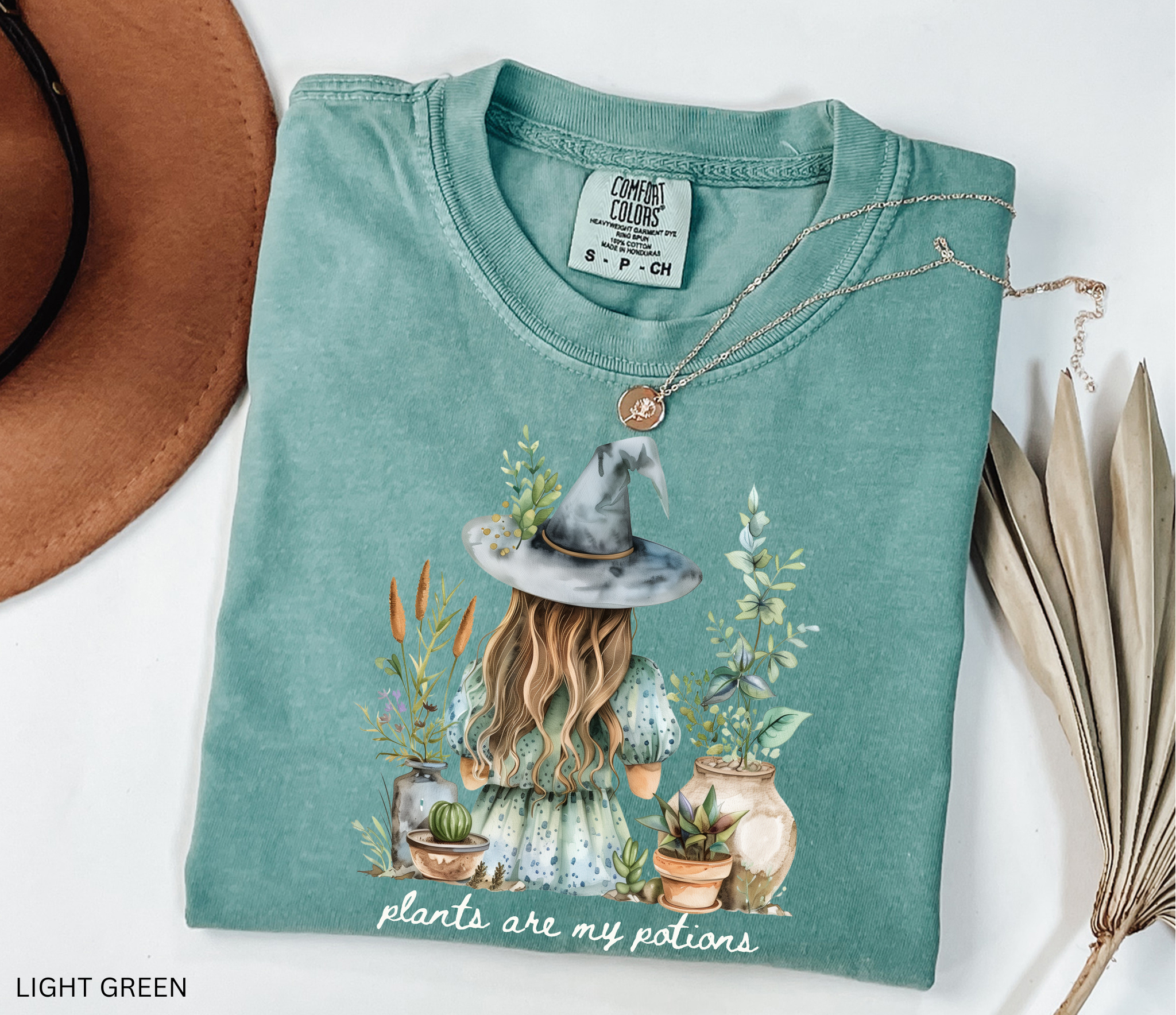 Witchy plant lover t-shirt with illustration of a witch in a hat surrounded by herbs and potted plants. Cottagecore gardening and green witch aesthetic.