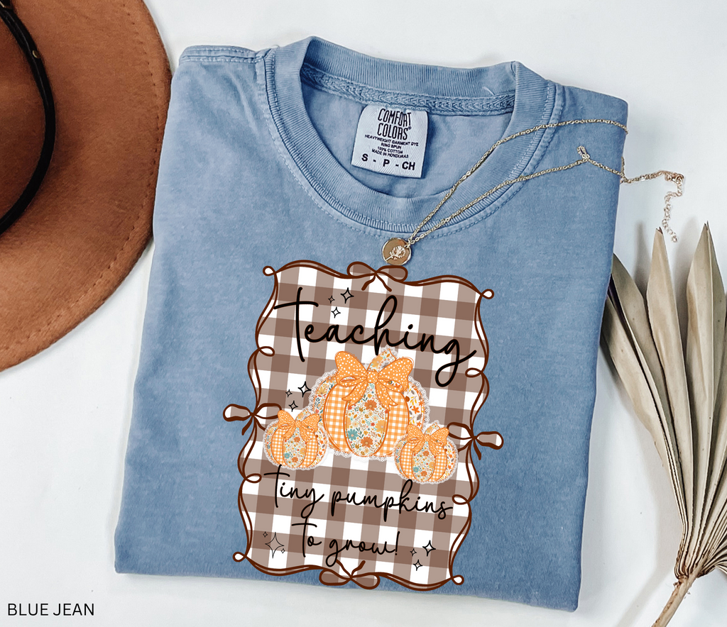 Brown gingham background Halloween teacher shirt with three floral orange pumpkins tied in bows and the phrase “Teaching Tiny Pumpkins to Grow.”