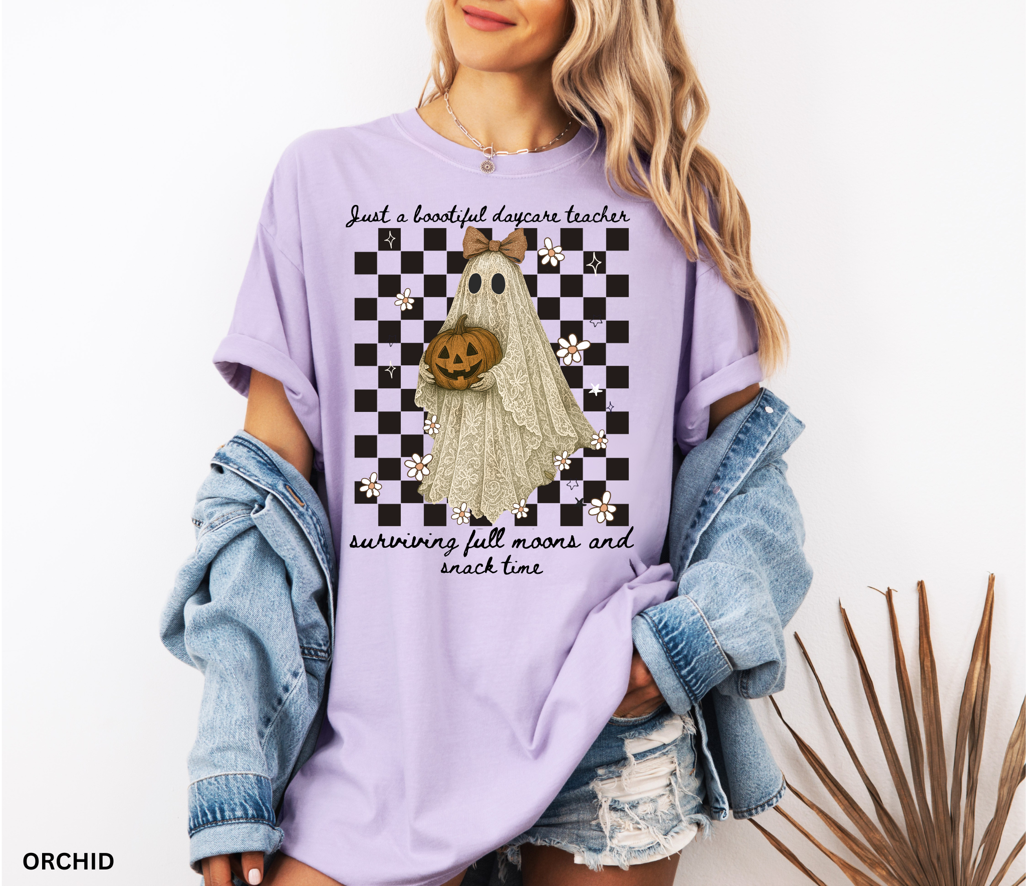 Daycare teacher Halloween shirt featuring a lace-patterned ghost holding a jack-o’-lantern pumpkin, wearing a bow, with flowers and stars on a checkered background.