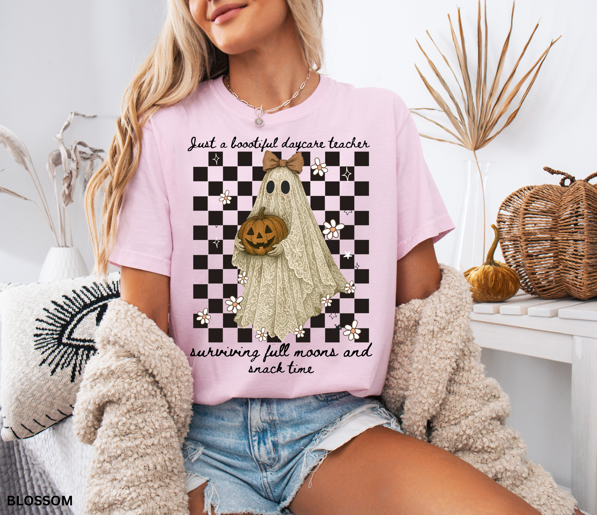 Daycare teacher Halloween shirt featuring a lace-patterned ghost holding a jack-o’-lantern pumpkin, wearing a bow, with flowers and stars on a checkered background.