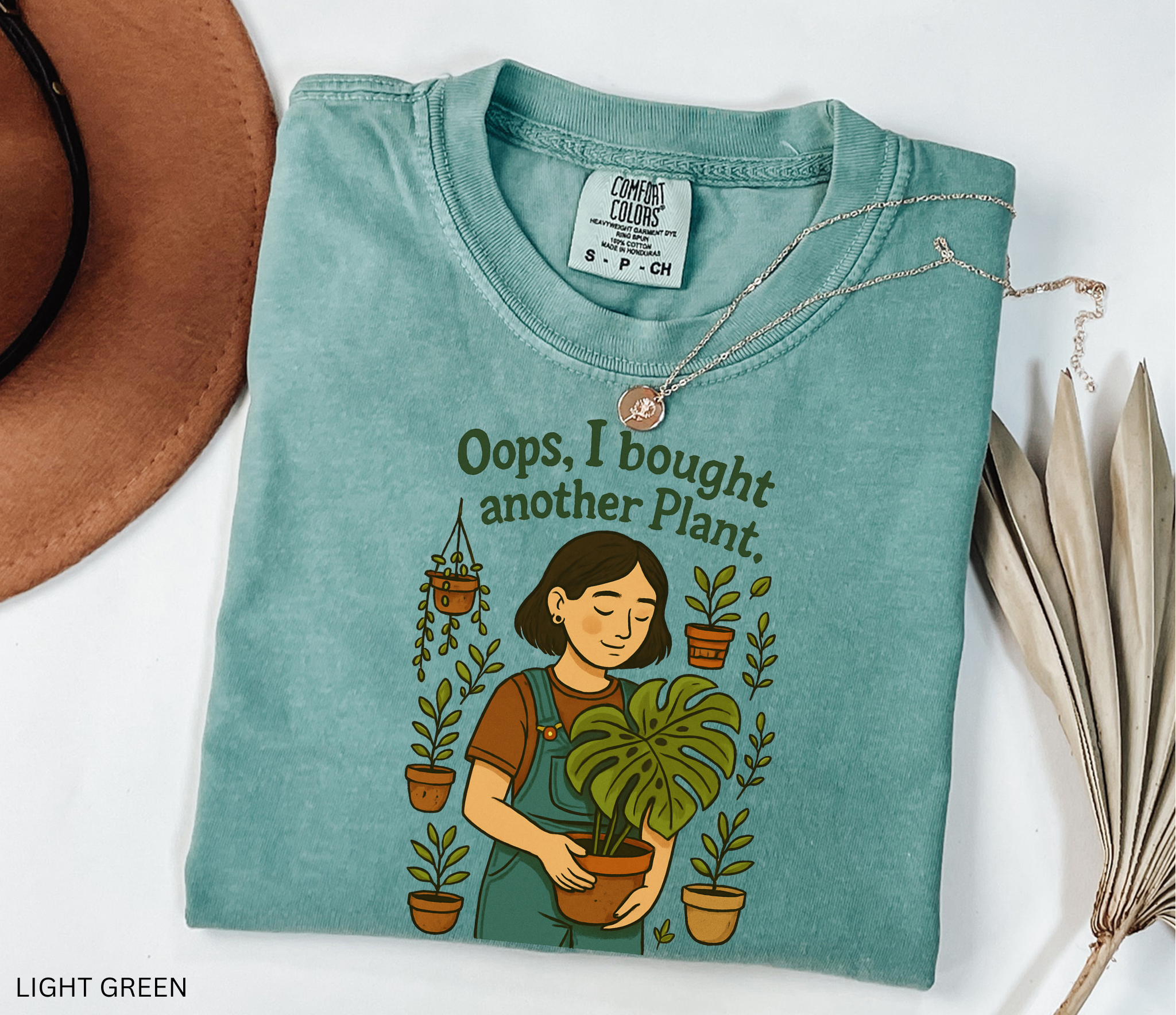 Gardening shirt with a woman in overalls holding a monstera plant, surrounded by potted plants, with the phrase “Oops, I Bought Another Plant.”