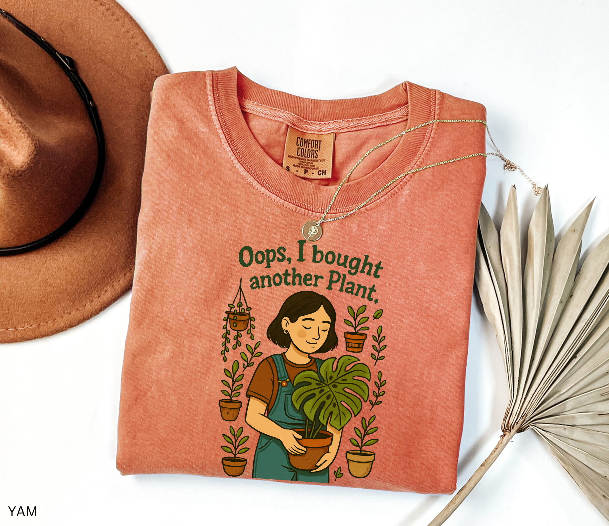Gardening shirt with a woman in overalls holding a monstera plant, surrounded by potted plants, with the phrase “Oops, I Bought Another Plant.”