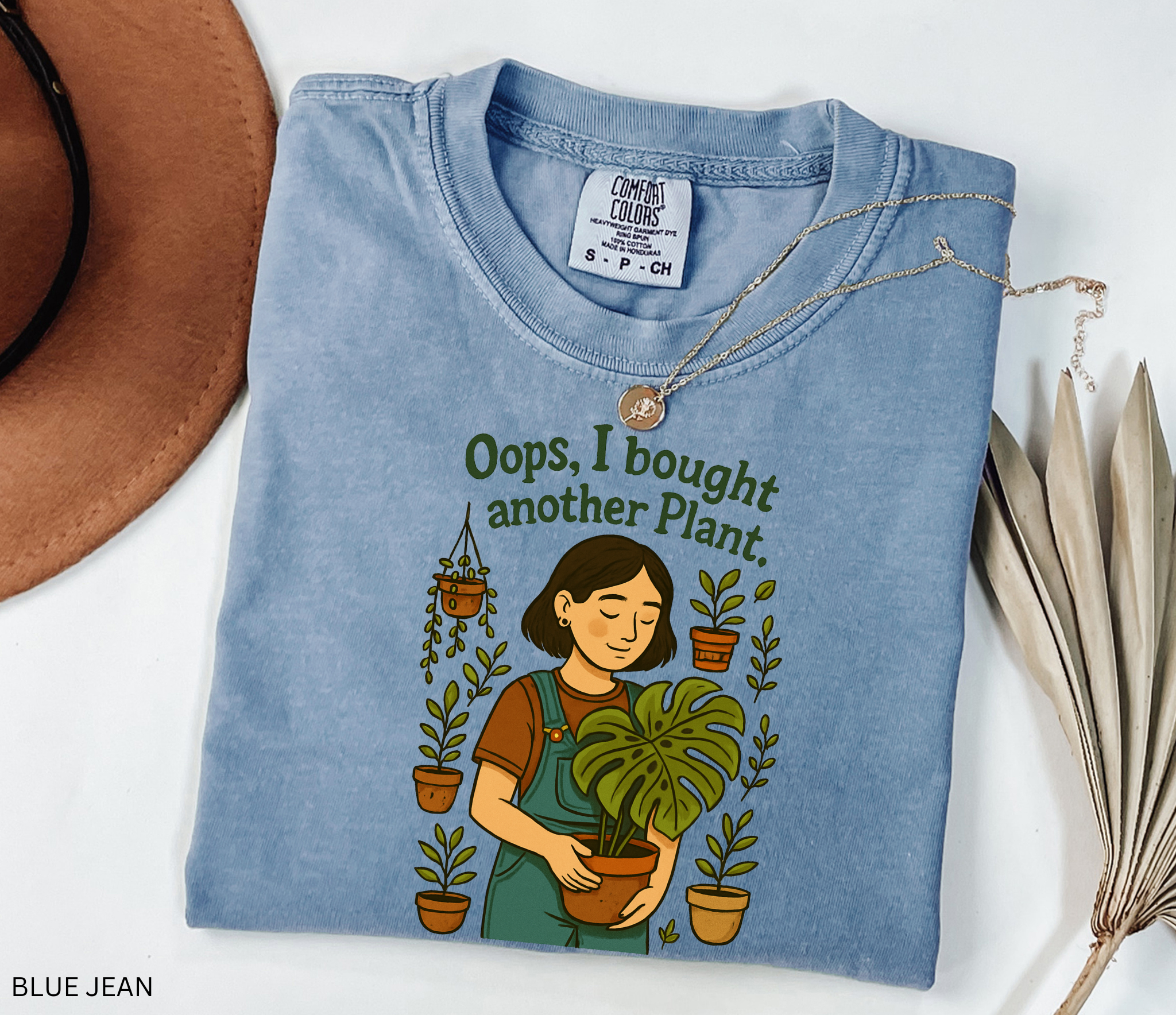 Gardening shirt with a woman in overalls holding a monstera plant, surrounded by potted plants, with the phrase “Oops, I Bought Another Plant.”