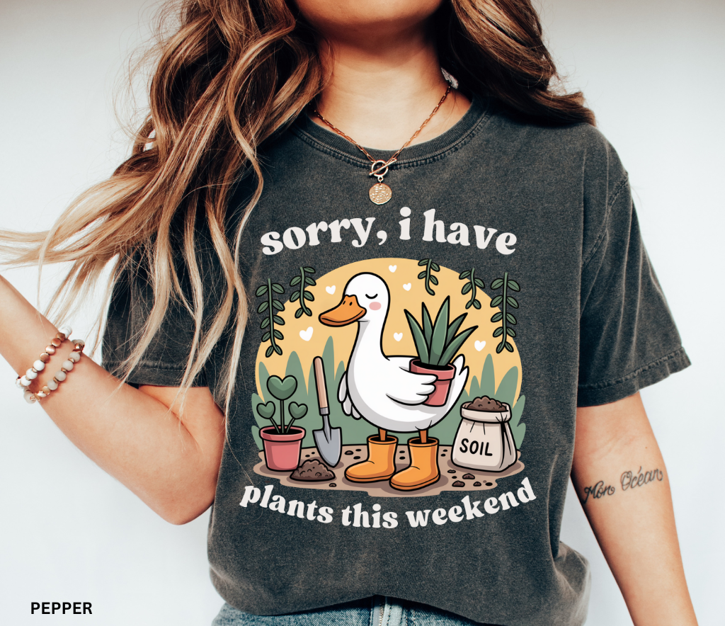 Gardening shirt with a cartoon goose in orange boots holding a potted plant, surrounded by soil, a shovel, and other plants in a cottagecore garden style.