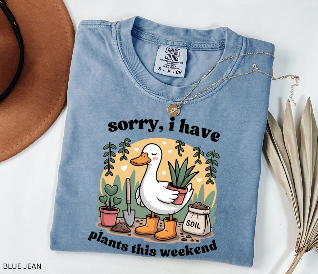 Gardening shirt with a cartoon goose in orange boots holding a potted plant, surrounded by soil, a shovel, and other plants in a cottagecore garden style.