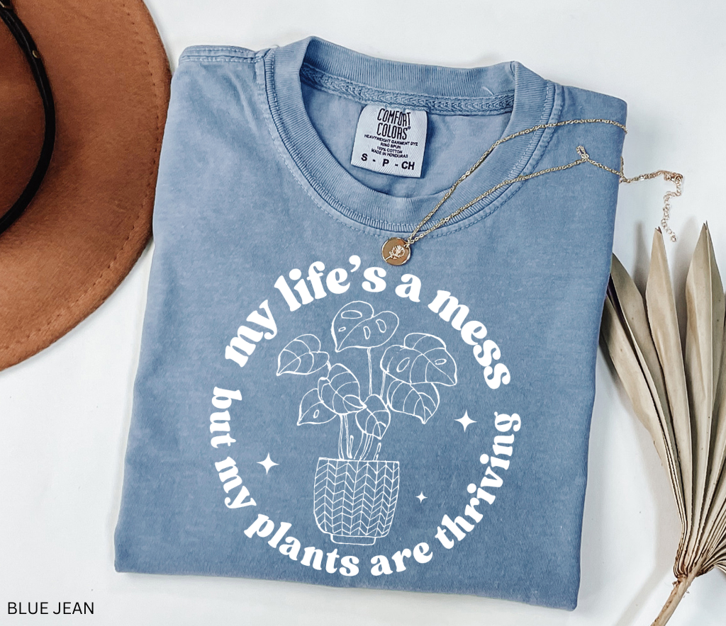 Black shirt with white line-art potted monstera plant and the phrase My Life’s a Mess But My Plants Are Thriving in bold rounded font.