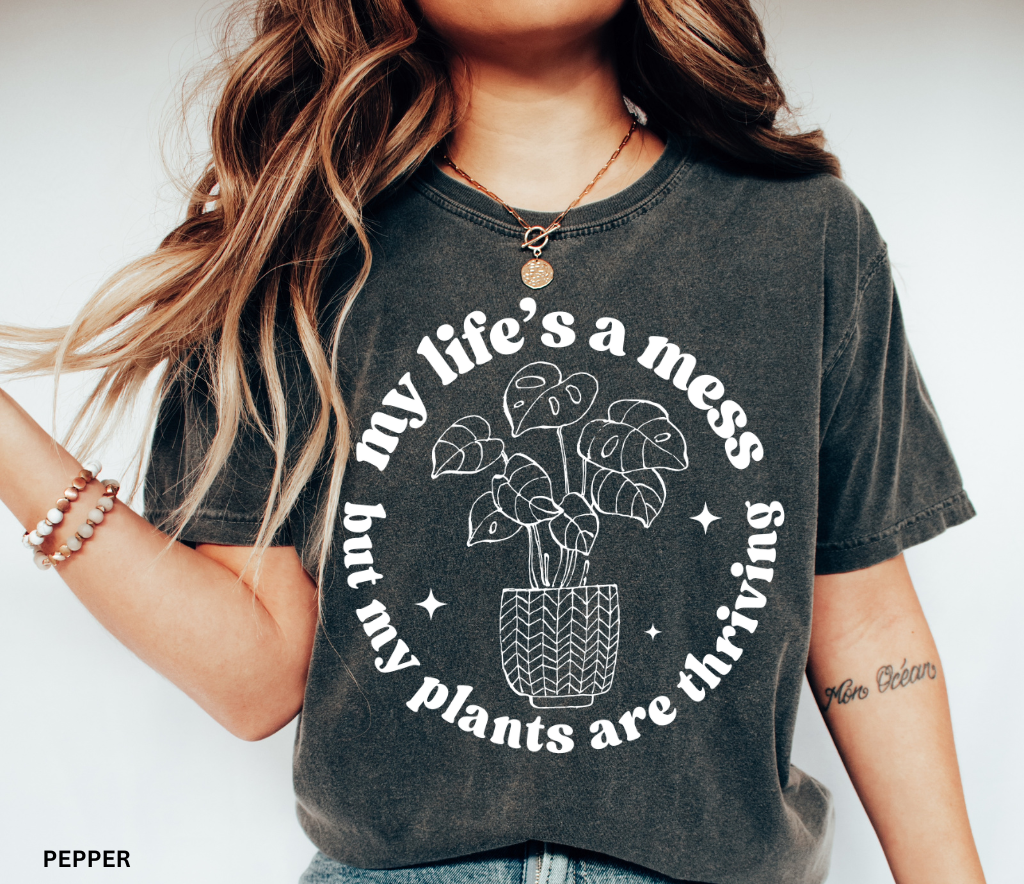 Black shirt with white line-art potted monstera plant and the phrase My Life’s a Mess But My Plants Are Thriving in bold rounded font.
