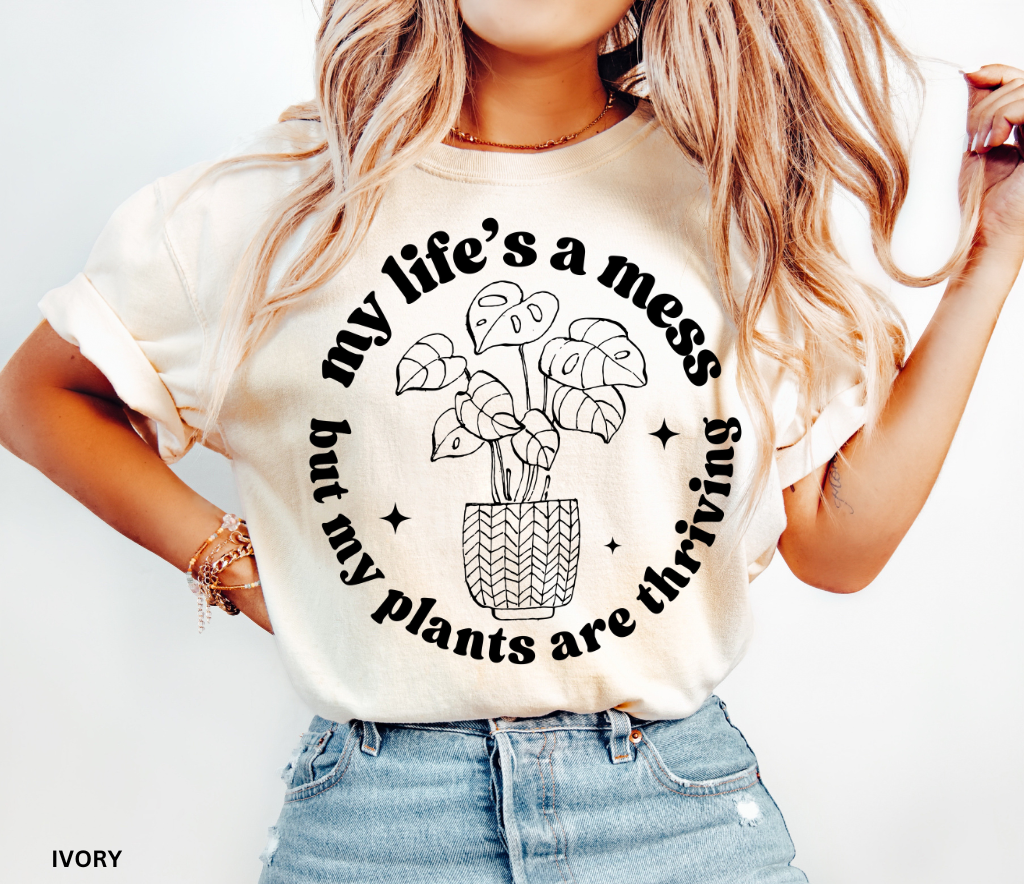 Black shirt with white line-art potted monstera plant and the phrase My Life’s a Mess But My Plants Are Thriving in bold rounded font.