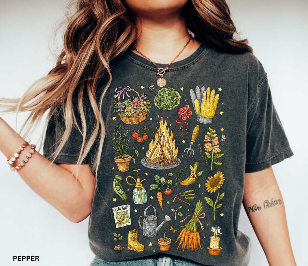 Black shirt with colorful cottagecore-style illustrations of plants, flowers, carrots, gardening tools, gloves, watering can, and a cozy campfire design.