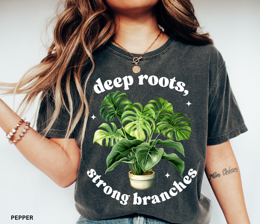 Black shirt with a large green monstera plant in a pot and the inspirational phrase Deep Roots, Strong Branches in white text.