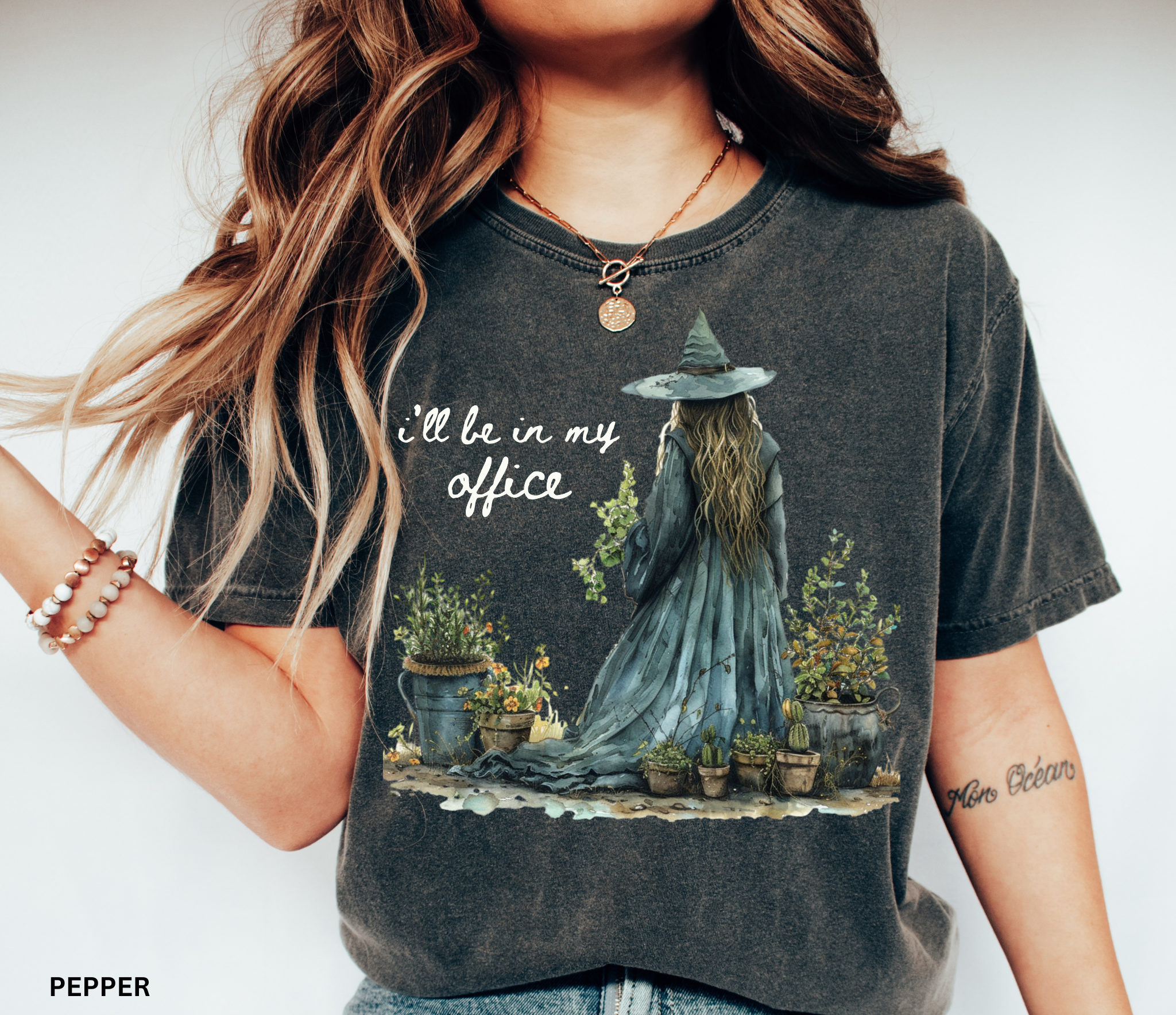 Watercolor-style illustration of a witch in a long flowing dress and hat, tending to potted plants and herbs, with the phrase “I’ll be in my office” on a cottagecore t-shirt.