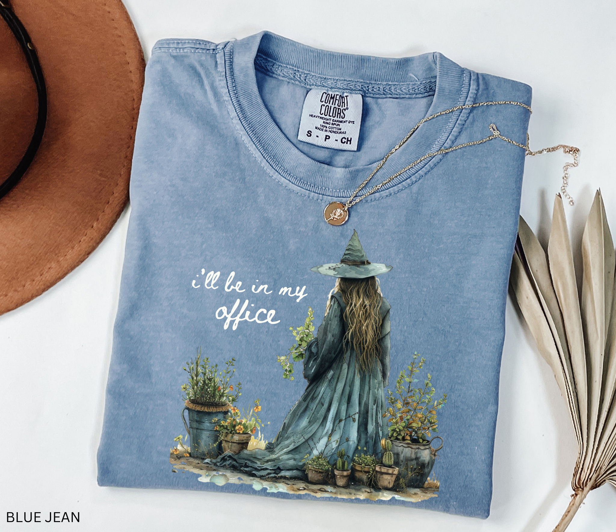 Watercolor-style illustration of a witch in a long flowing dress and hat, tending to potted plants and herbs, with the phrase “I’ll be in my office” on a cottagecore t-shirt.