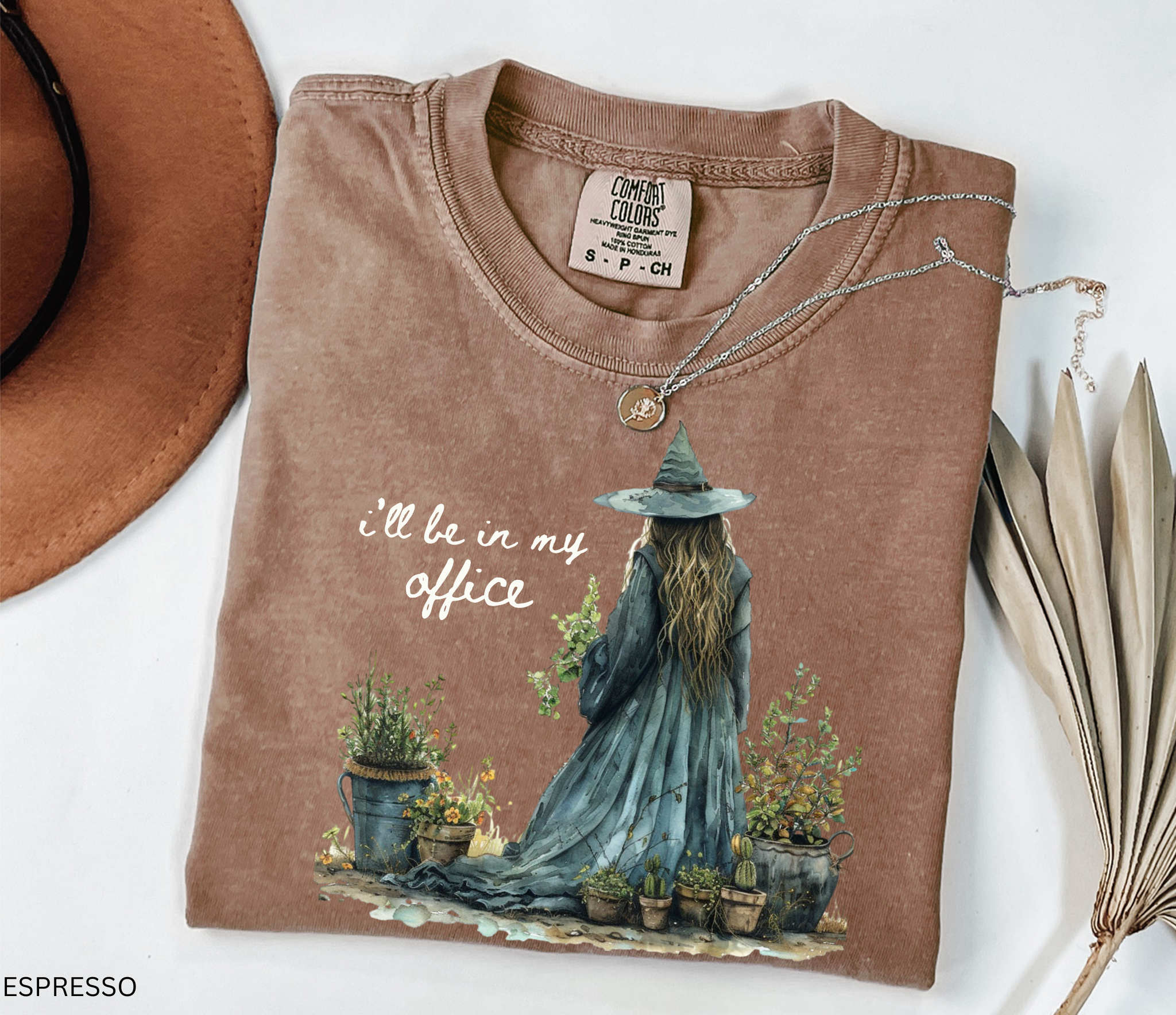 Watercolor-style illustration of a witch in a long flowing dress and hat, tending to potted plants and herbs, with the phrase “I’ll be in my office” on a cottagecore t-shirt.