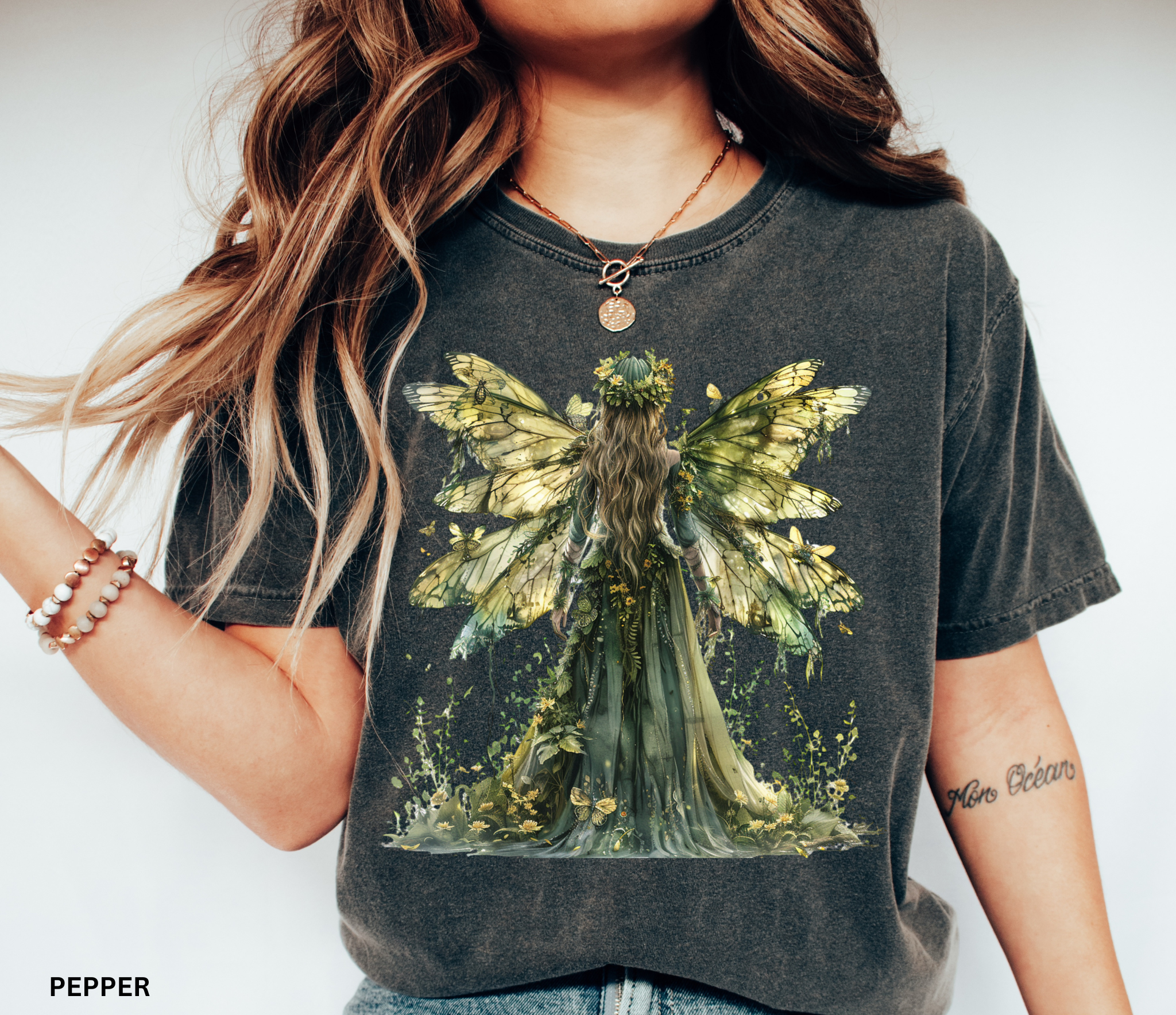 Watercolor-style illustration of a fairy with large golden butterfly wings and a flowing green gown, surrounded by flowers and plants, printed on a cottagecore t-shirt.