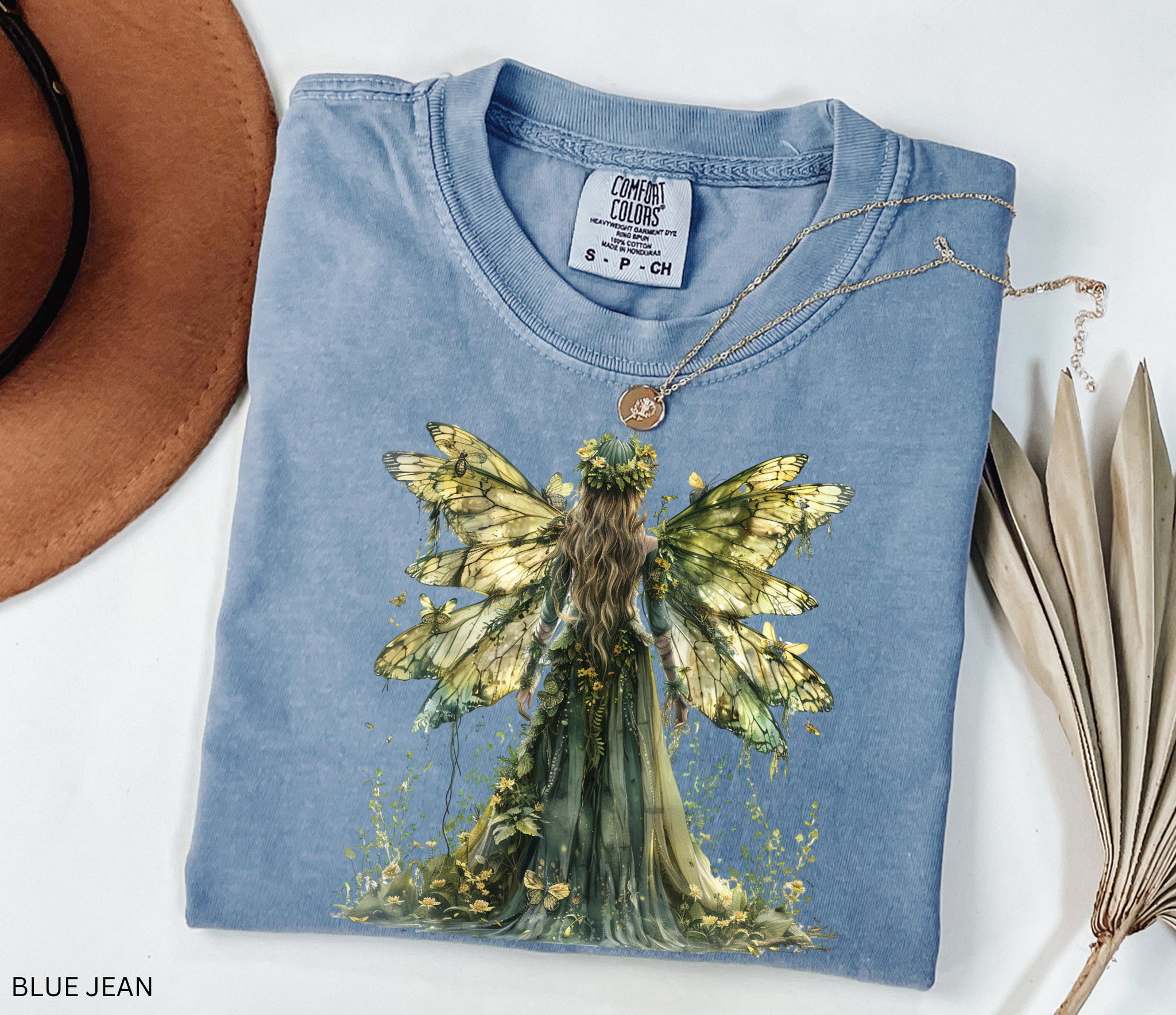 Watercolor-style illustration of a fairy with large golden butterfly wings and a flowing green gown, surrounded by flowers and plants, printed on a cottagecore t-shirt.