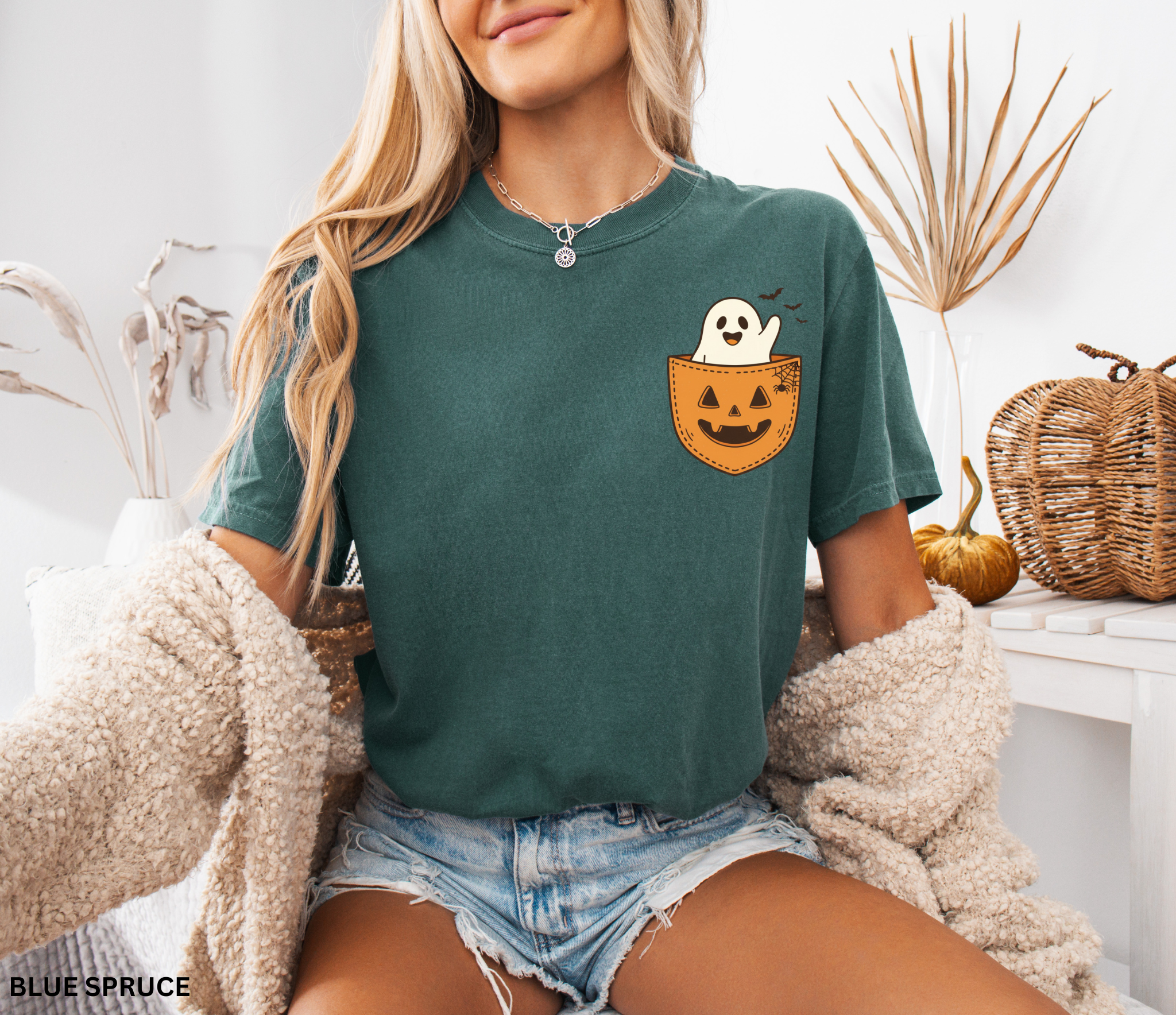 Cute ghost peeking out of a jack-o’-lantern pocket with bats and spiderweb detail on a Halloween t-shirt.
