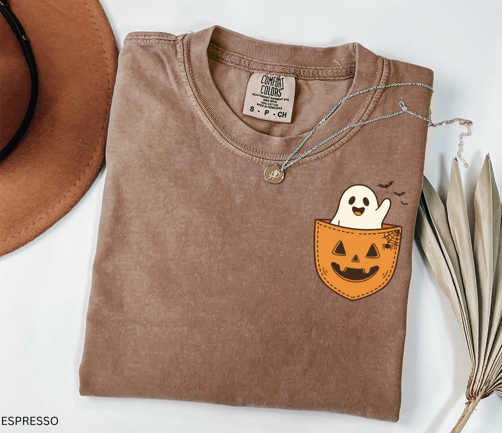 Cute ghost peeking out of a jack-o’-lantern pocket with bats and spiderweb detail on a Halloween t-shirt.