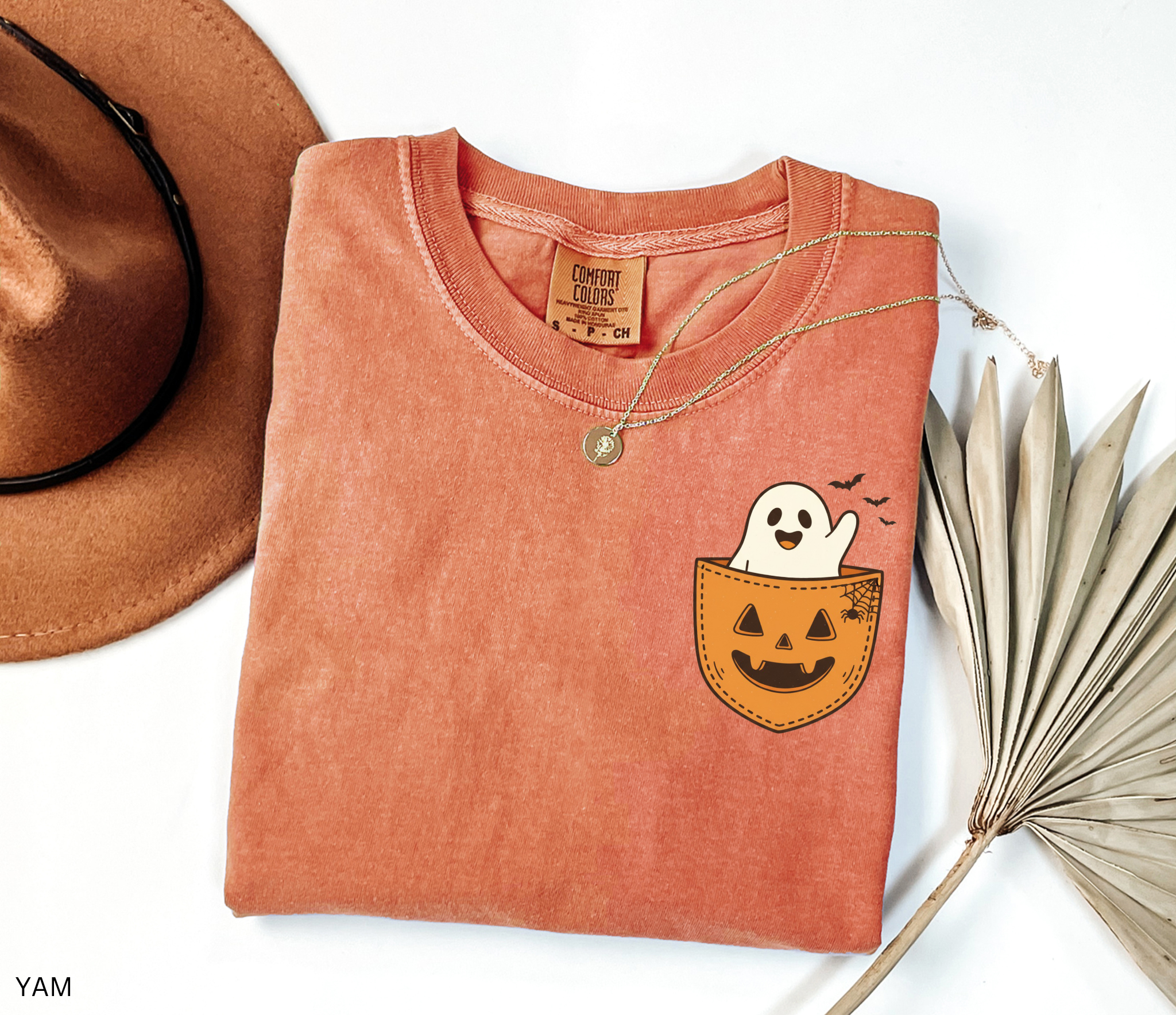 Cute ghost peeking out of a jack-o’-lantern pocket with bats and spiderweb detail on a Halloween t-shirt.
