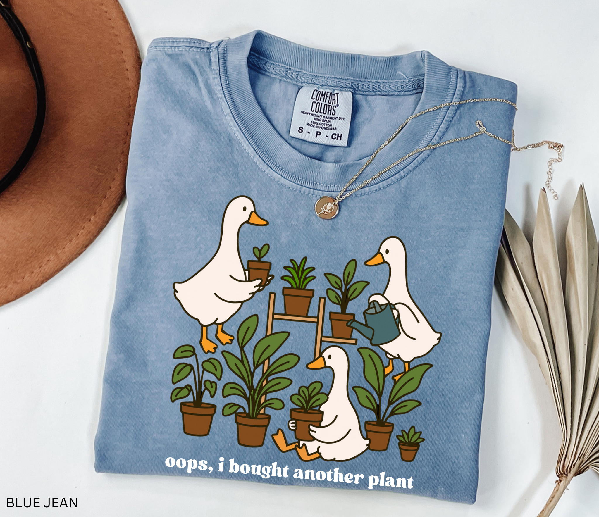 Funny gardening shirt with geese holding potted plants and watering cans, surrounded by greenery, with the phrase “Oops, I Bought Another Plant.”