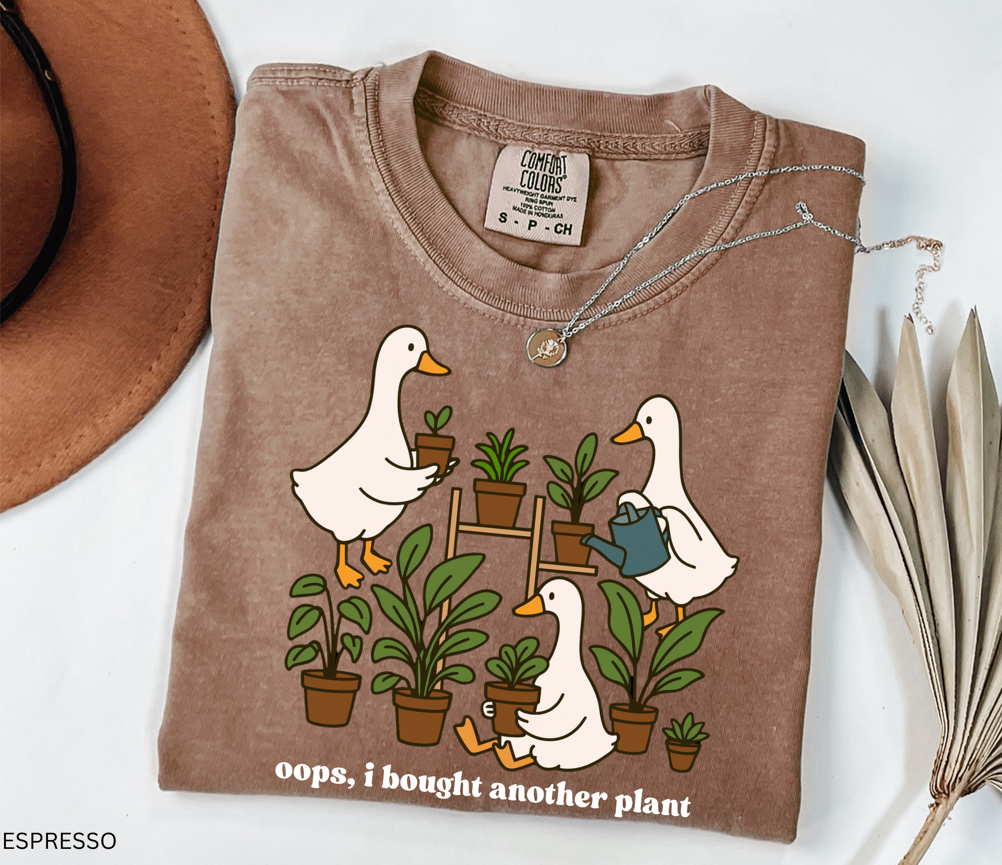 Funny gardening shirt with geese holding potted plants and watering cans, surrounded by greenery, with the phrase “Oops, I Bought Another Plant.”