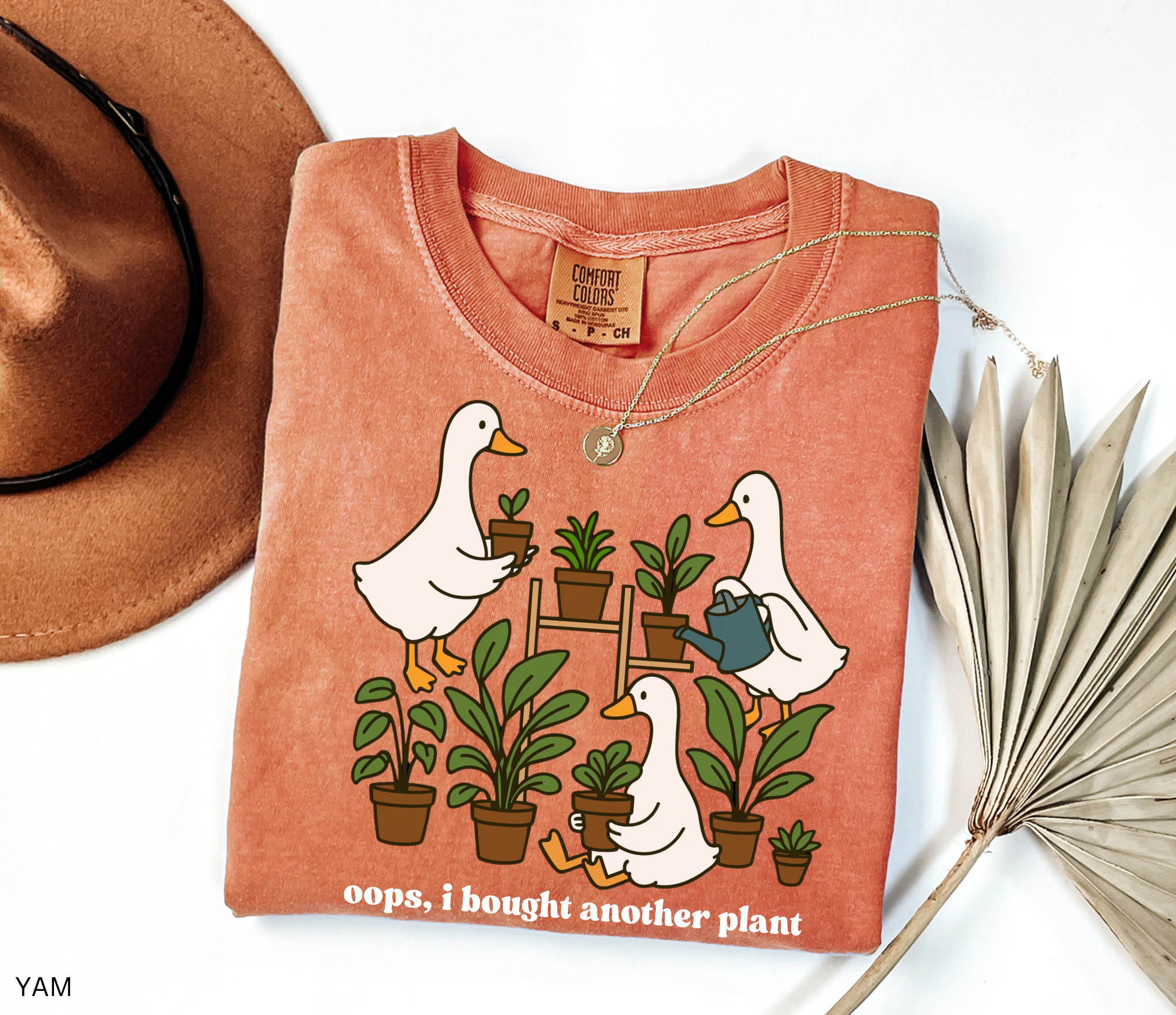 Funny gardening shirt with geese holding potted plants and watering cans, surrounded by greenery, with the phrase “Oops, I Bought Another Plant.”
