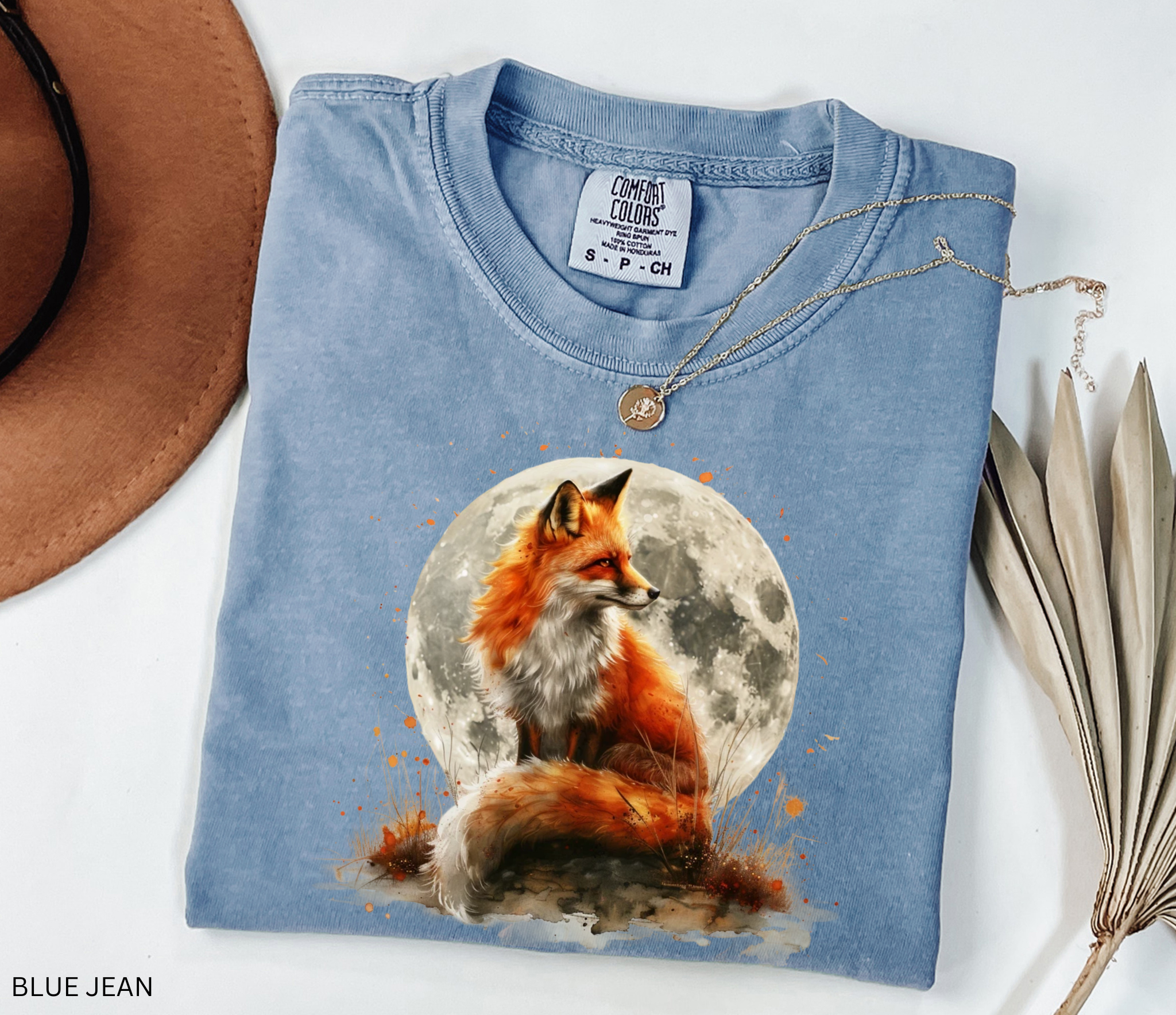 Watercolor-style illustration of a fox sitting beneath a glowing full moon, surrounded by grass and earthy details on a cottagecore shirt.