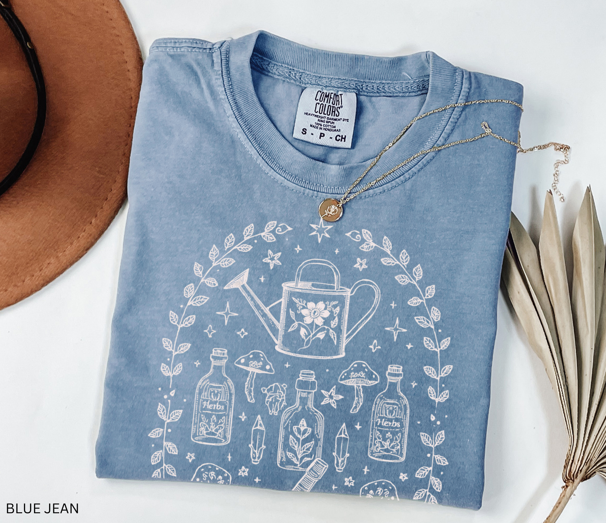 Hand-drawn botanical witch t-shirt featuring vintage herb bottles, mushrooms, crystals, and watering can – mystical garden witch aesthetic design on Comfort Colors tee.