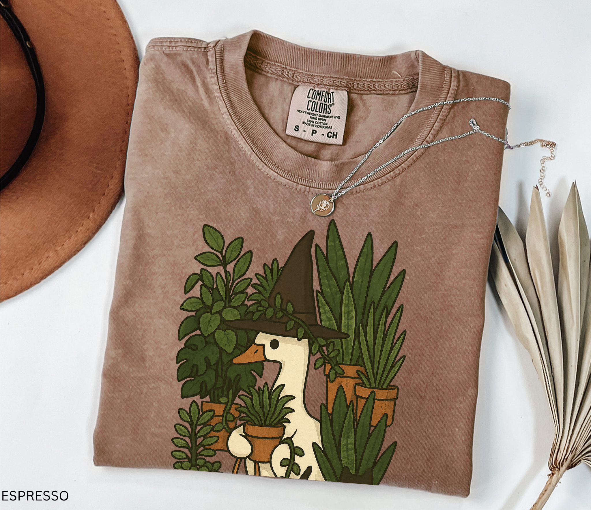 Goose wearing a witch hat surrounded by plants with a black cat companion — cozy cottagecore witchy design on Comfort Colors shirt.