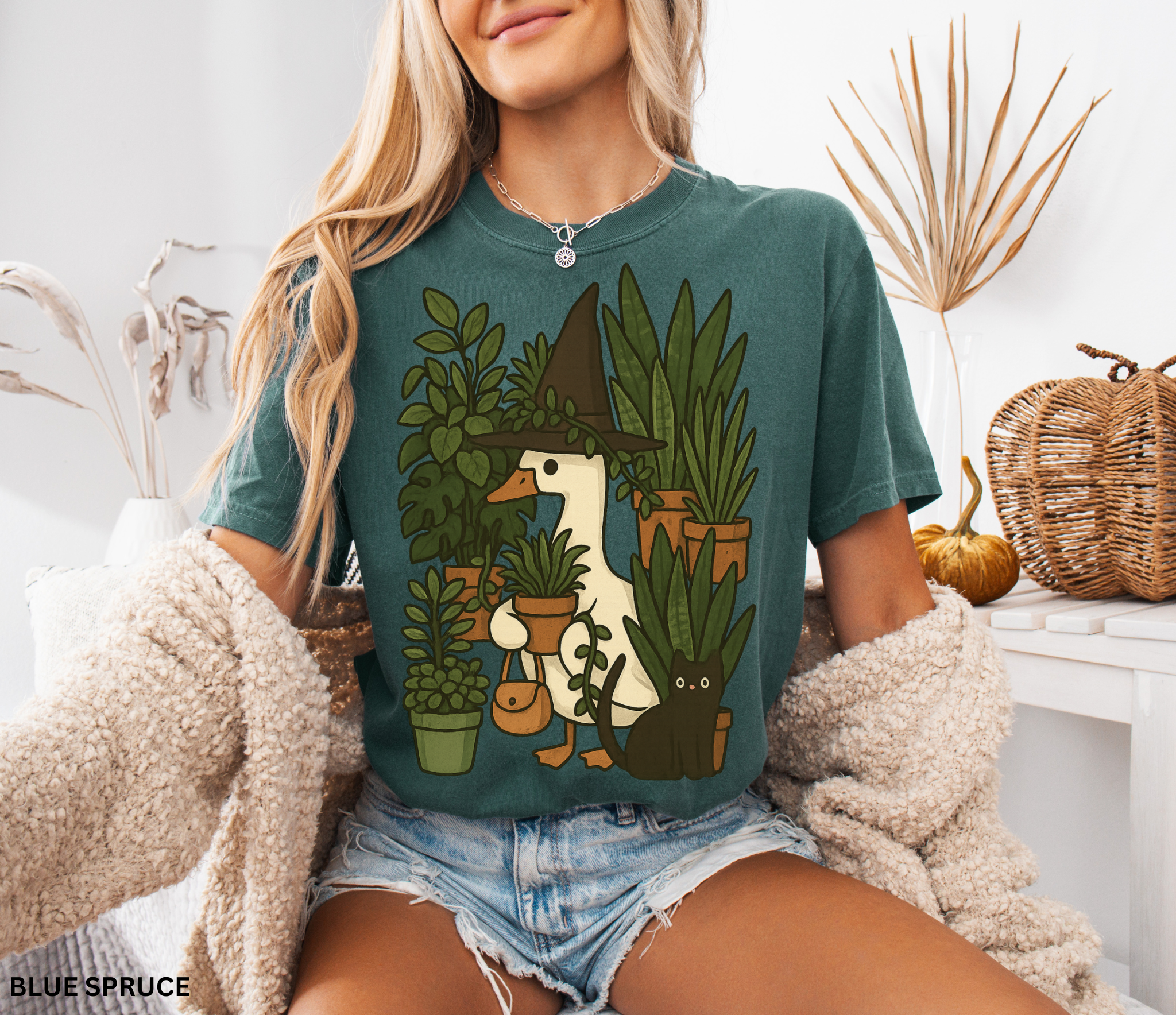 Goose wearing a witch hat surrounded by plants with a black cat companion — cozy cottagecore witchy design on Comfort Colors shirt.